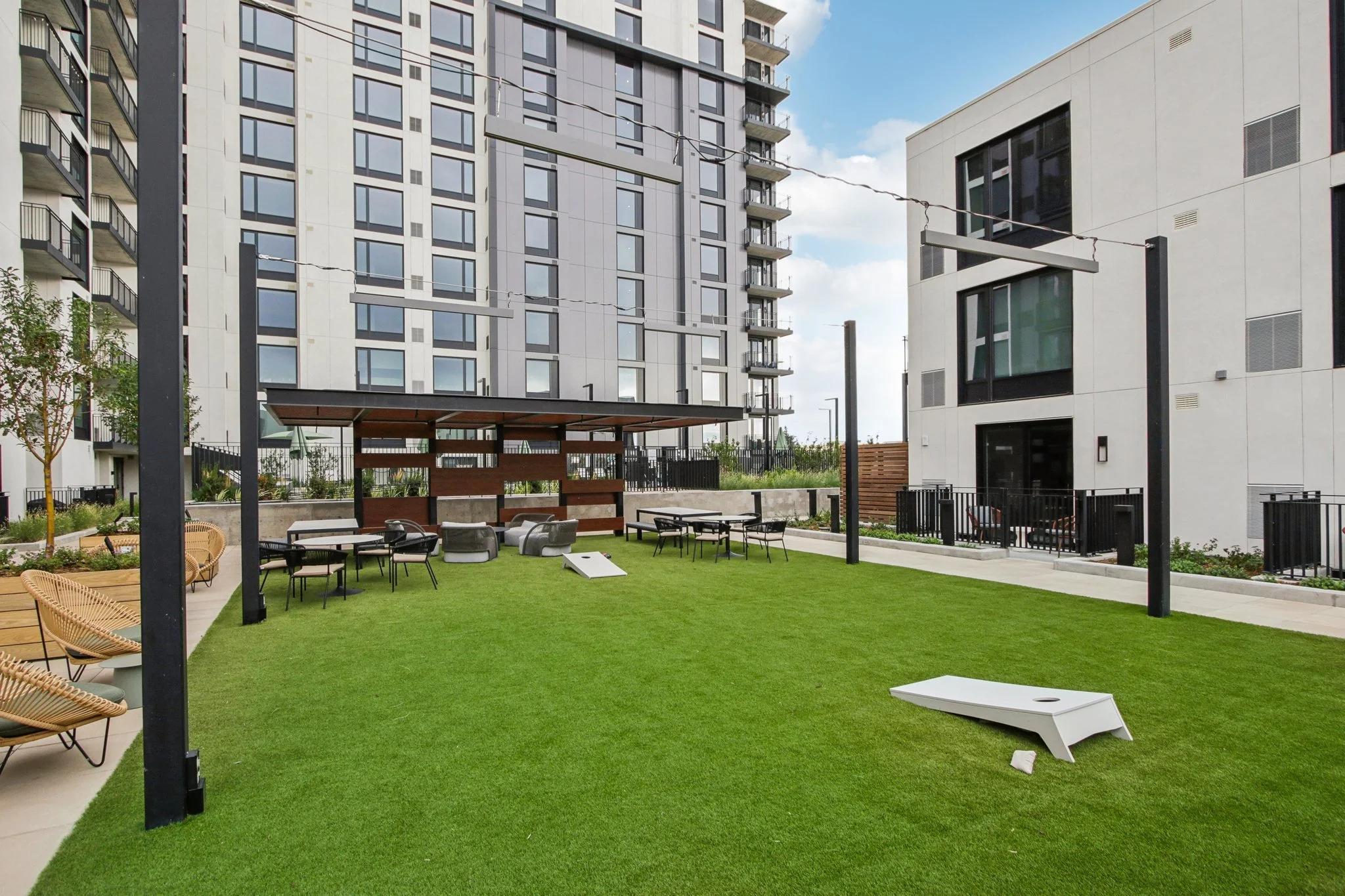 An outdoor communal space with a green artificial grass lawn, surrounded by modern multi-story apartment buildings. The area includes tables and chairs, a covered seating area with a wooden slatted wall, a cornhole game, string lights overhead, and a