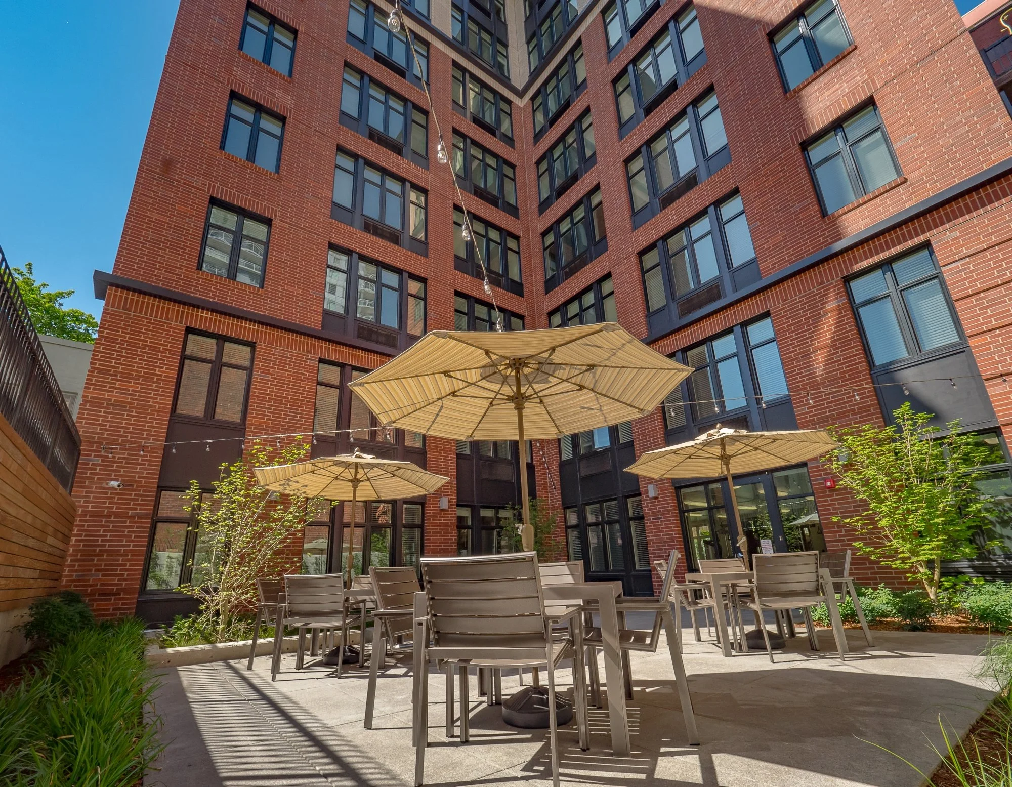 Outdoor courtyard with tables and umbrellas in front of a red brick apartment building.