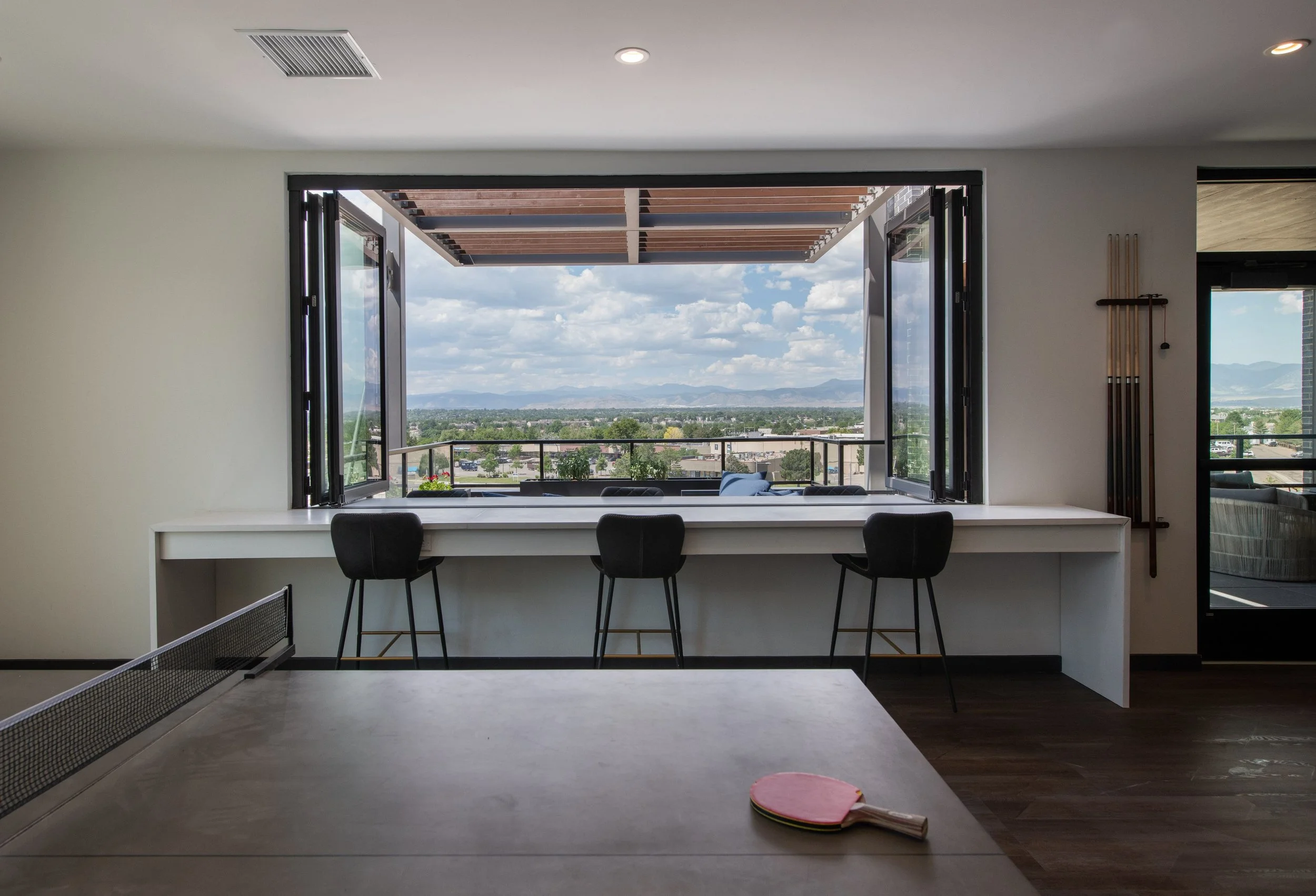 Indoor room with a large open window overlooking a city skyline and mountains in the distance. There are three barstools along a white counter beneath the window, and a ping pong paddle and ball on a table in the foreground.