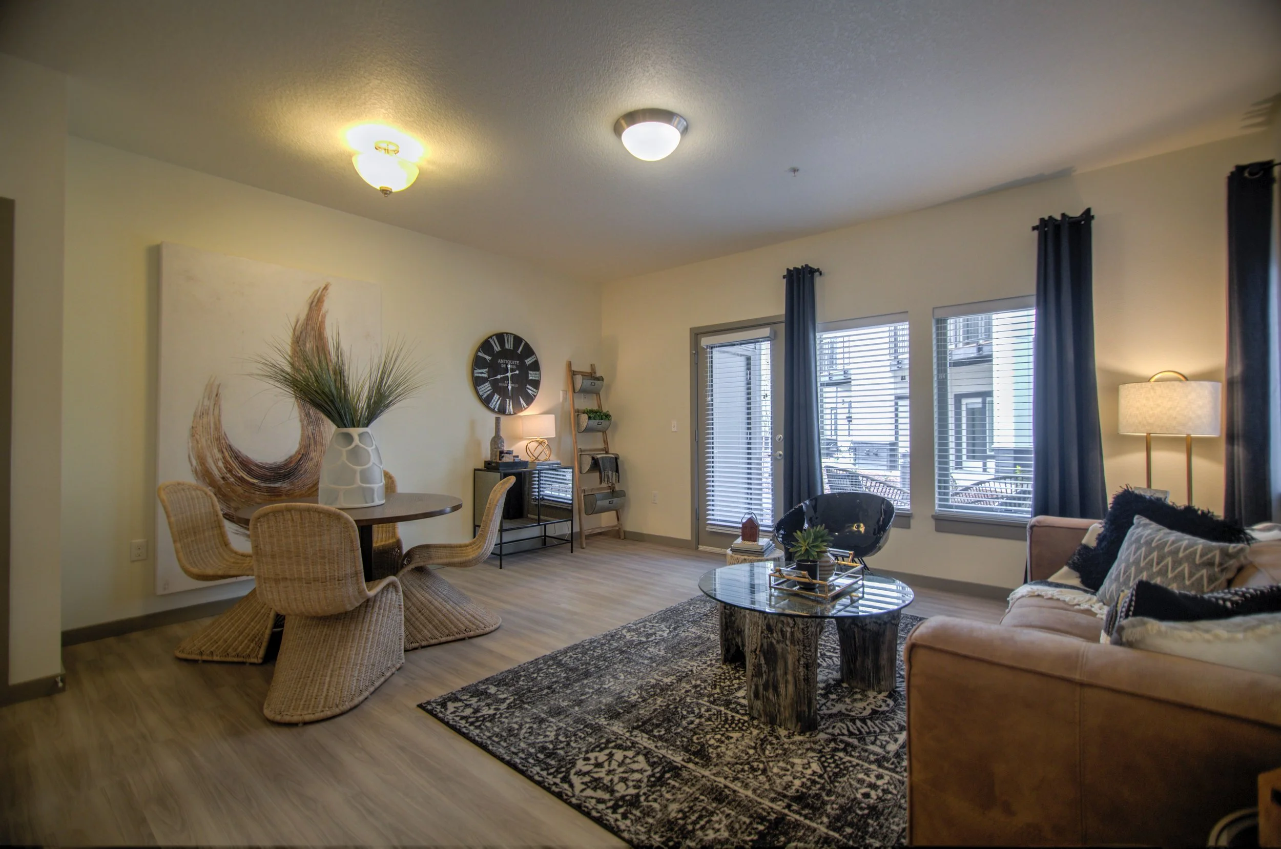 Living room with a beige sofa, black round chair, glass coffee table, and large windows with blinds and blue curtains, decorated with artwork, plants, and lamps.