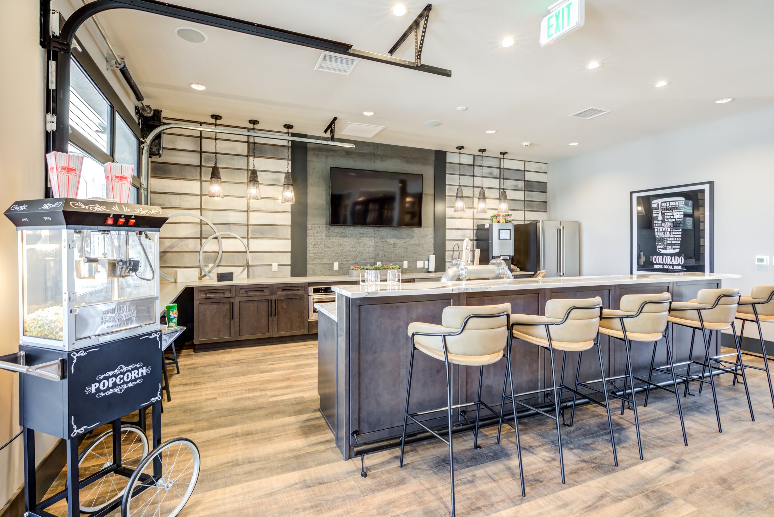 Modern coffee shop or cafe interior with a long bar counter, high chairs, popcorn machine, and kitchen area with hanging pendant lights and a TV on the wall.