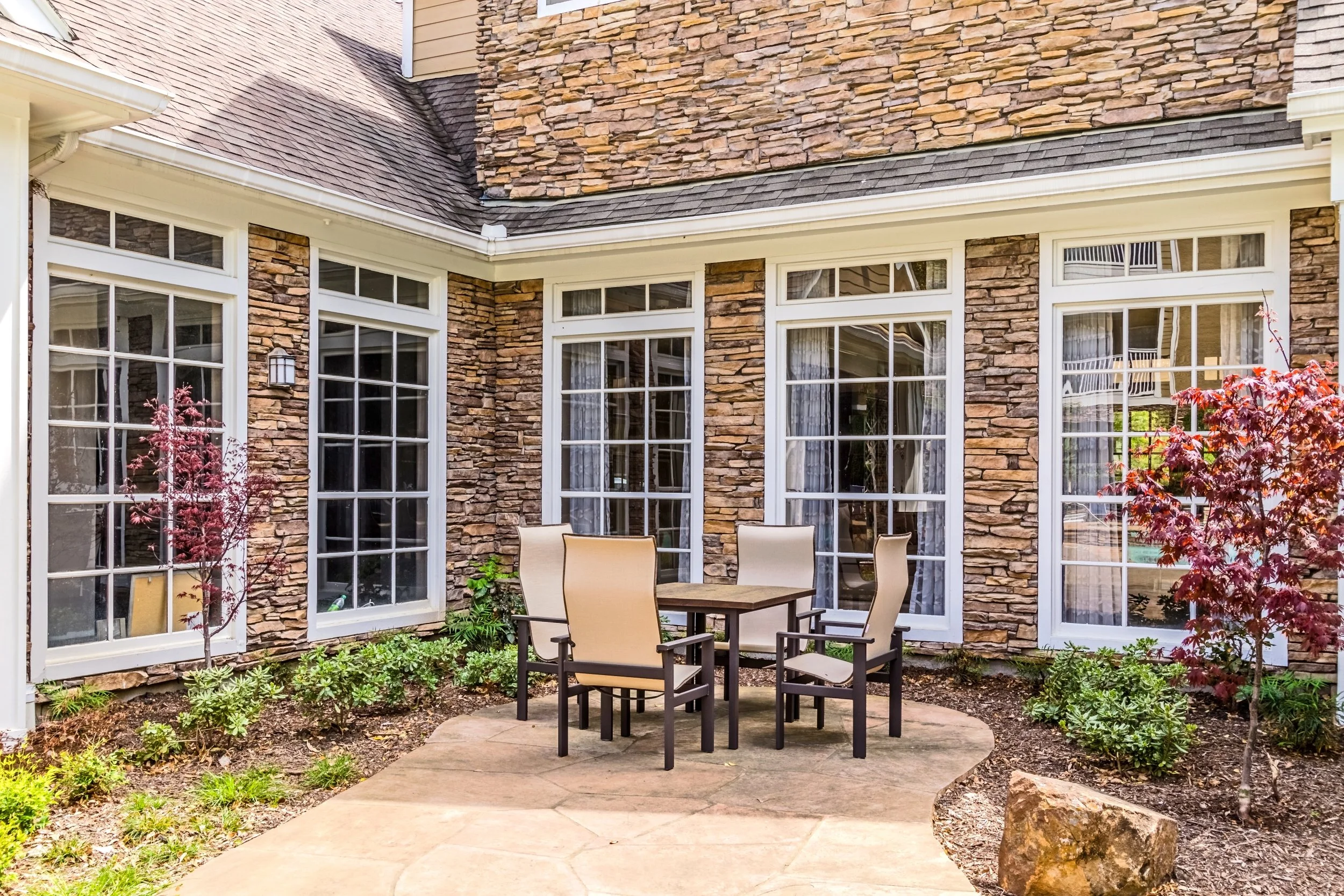 Outdoor patio with a table and four chairs, surrounded by house with large windows and stone siding, plants, and small trees.