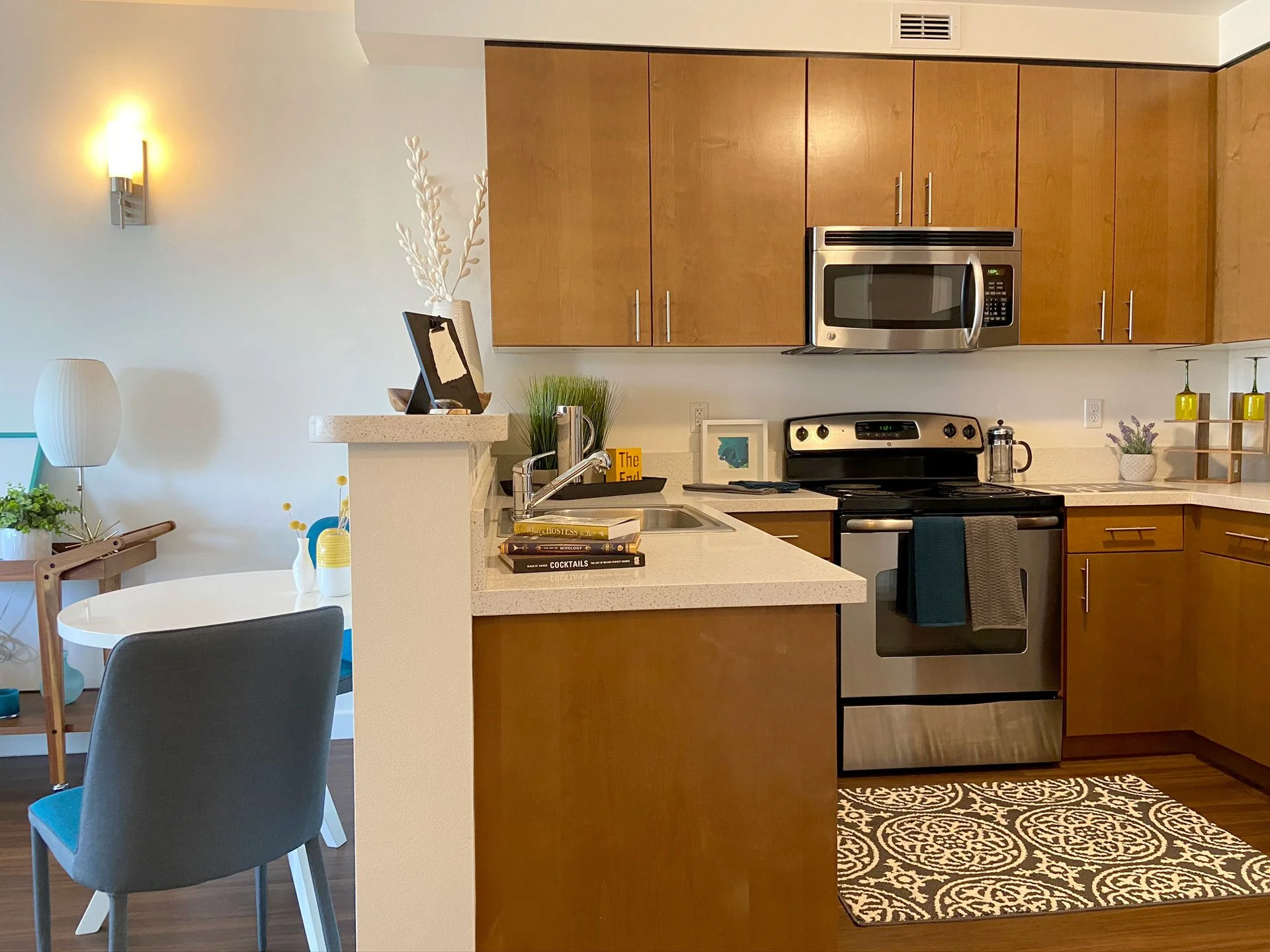 Modern kitchen with wooden cabinets, stainless steel appliances including microwave and oven, decorative plants, a small table with chairs, and a patterned rug on a wooden floor.