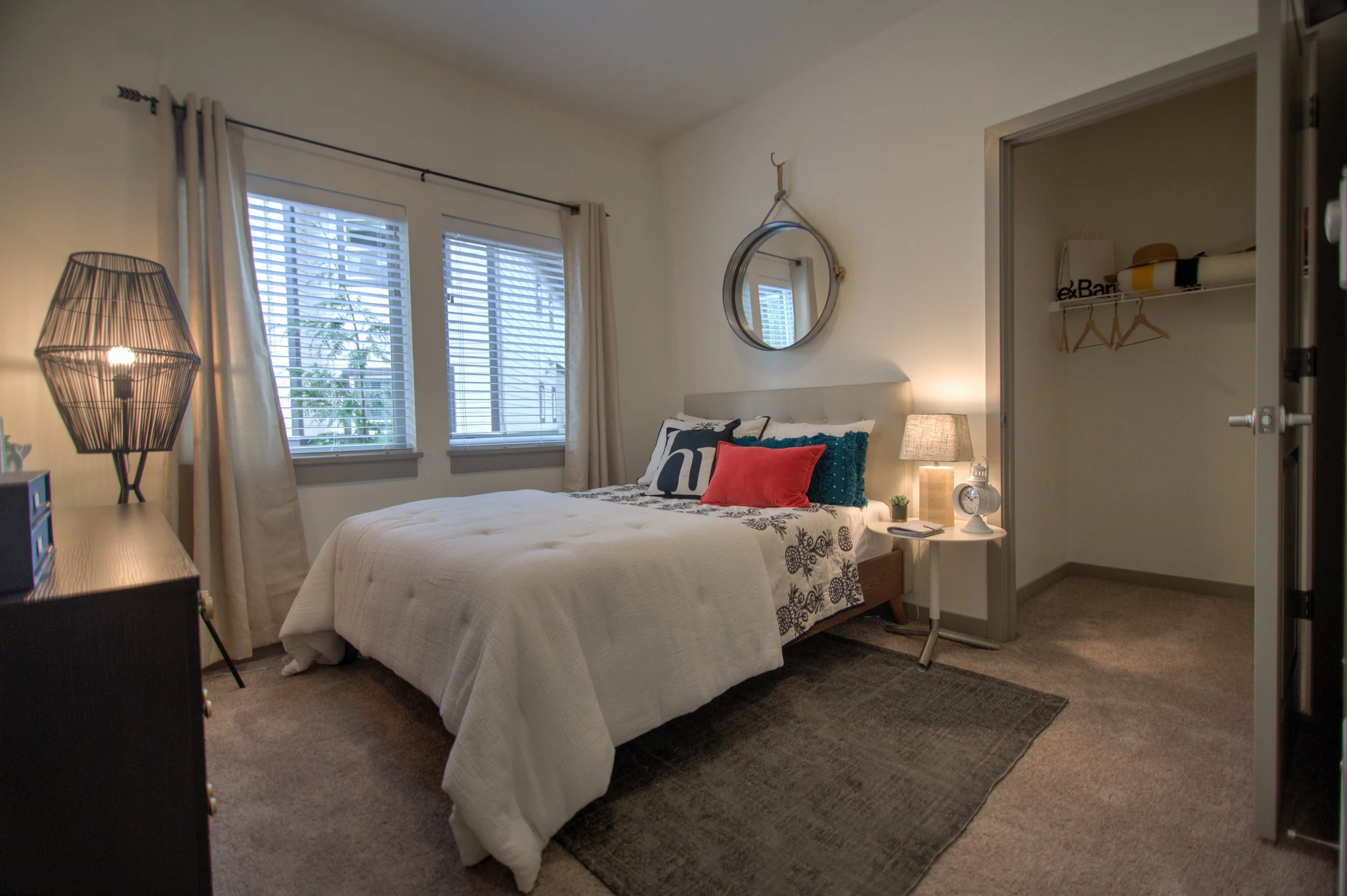 A cozy bedroom with a bed against the wall, three decorative pillows, a side table with a lamp, and a closet with open shelves and hangers.
