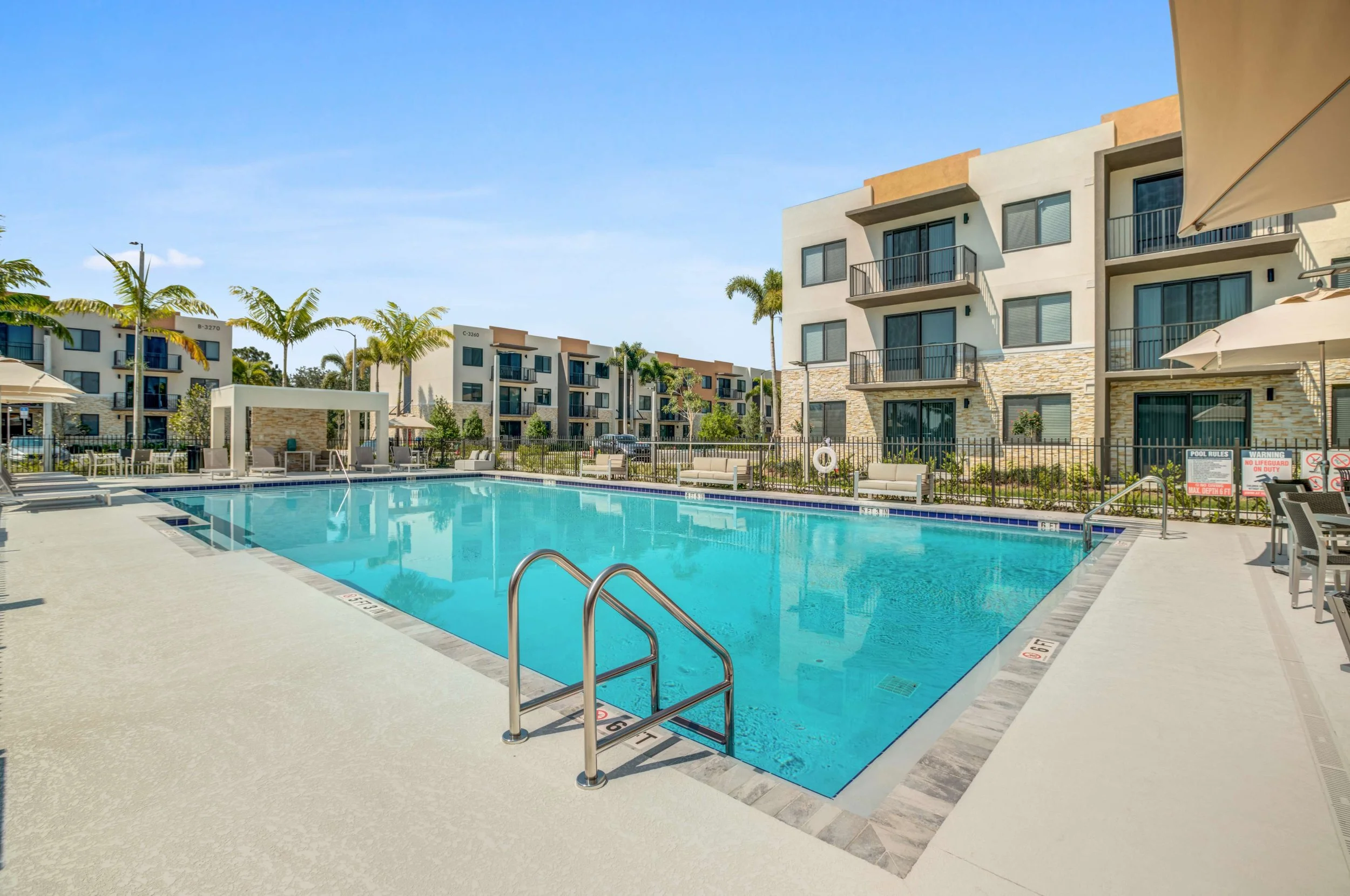 Outdoor swimming pool in a modern apartment complex with lounge chairs, umbrellas, and surrounding buildings with balconies, palm trees, and clear blue sky.