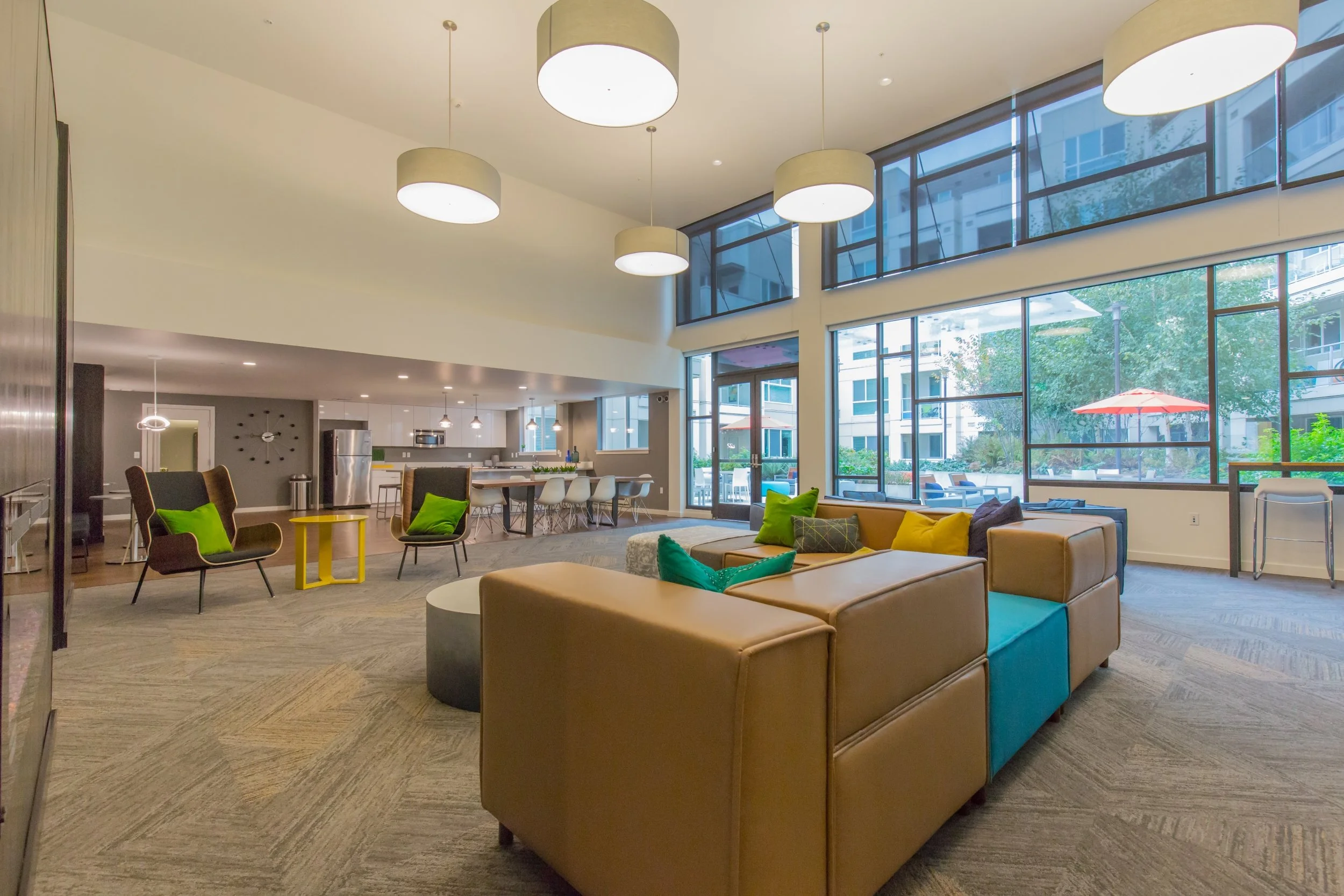 Modern apartment living room with large windows, beige sofa with colorful pillows, black armchairs with green cushions, and a kitchen area in the background.