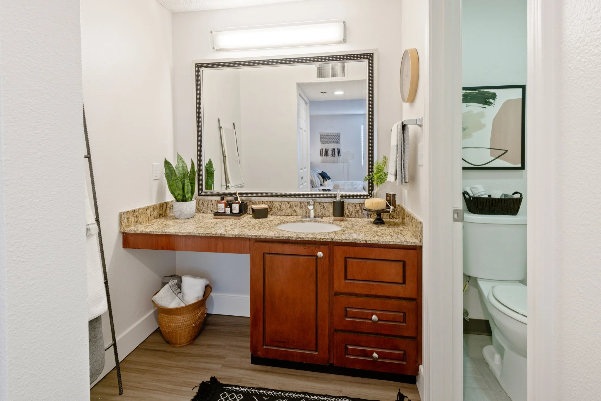 Bathroom vanity with a large mirror, a granite countertop, and wooden cabinets. Toilet is visible through an open door. Decorative items and plants on the countertop.