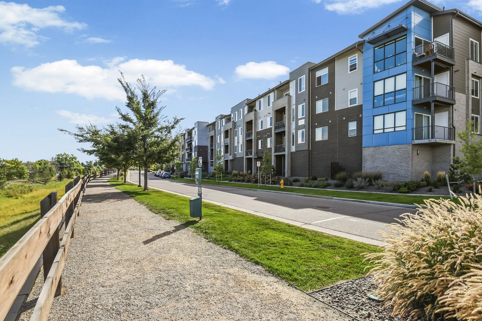 View of modern apartment building with balconies, alongside a sidewalk with trees and grass on a sunny day.