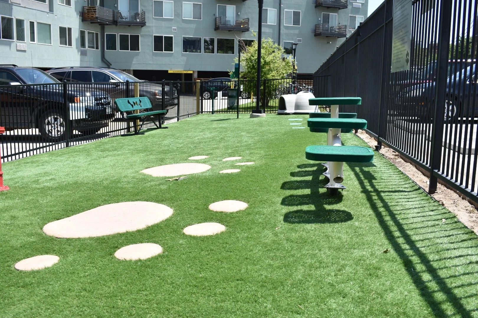 Outdoor dog park with artificial grass, rock-shaped stepping stones, green benches, picnic table, fenced enclosure, and parked cars in the background.