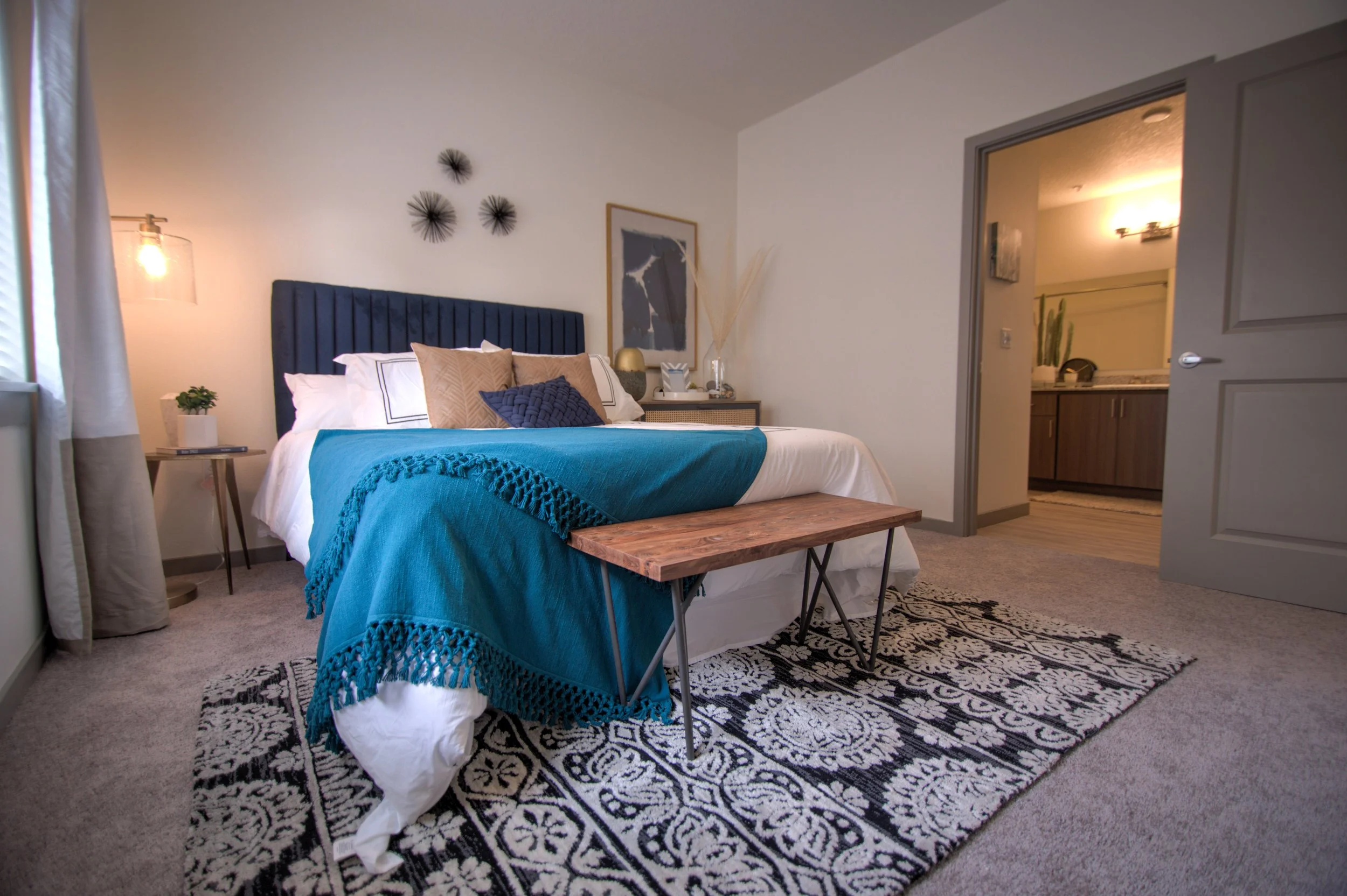 A bedroom with a king-sized bed, blue headboard, white bedding, and decorative pillows, with a blanket draped over the side. There is a wooden bench at the foot of the bed, a patterned area rug, and a nightstand with a plant and a lamp. An abstract a