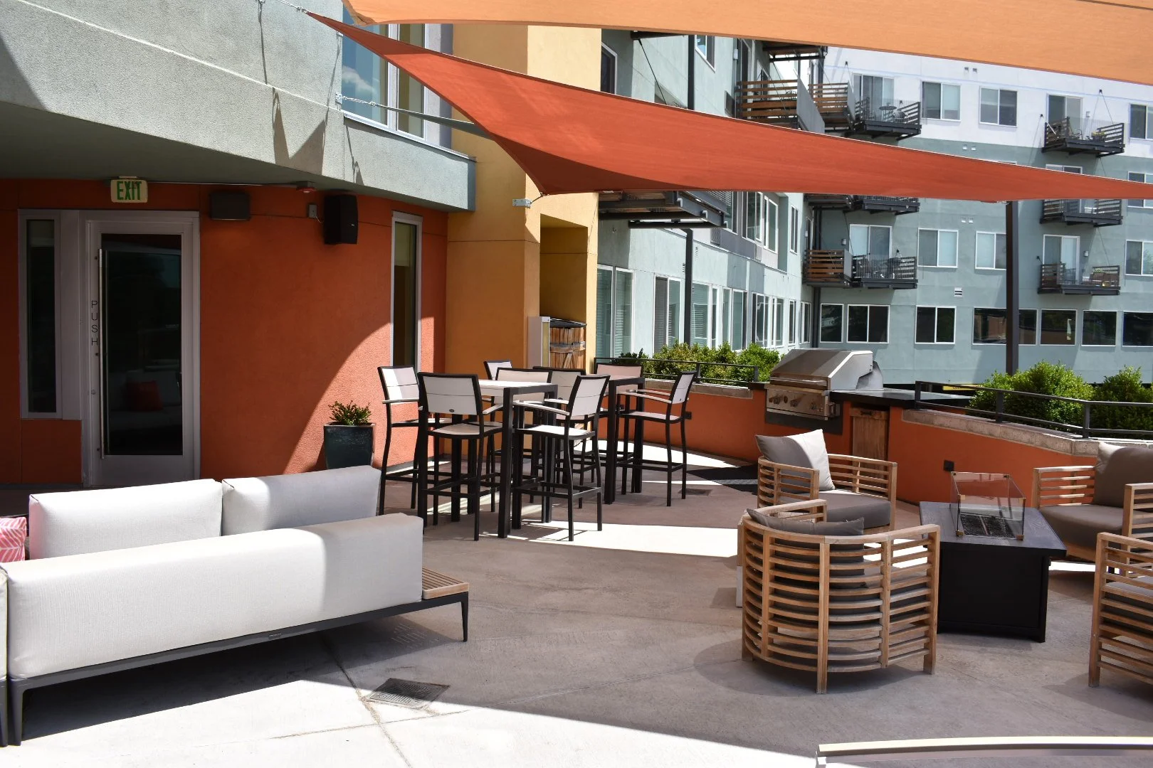 Outdoor patio with tables and chairs, sun shading, grill, and neighboring apartment building.