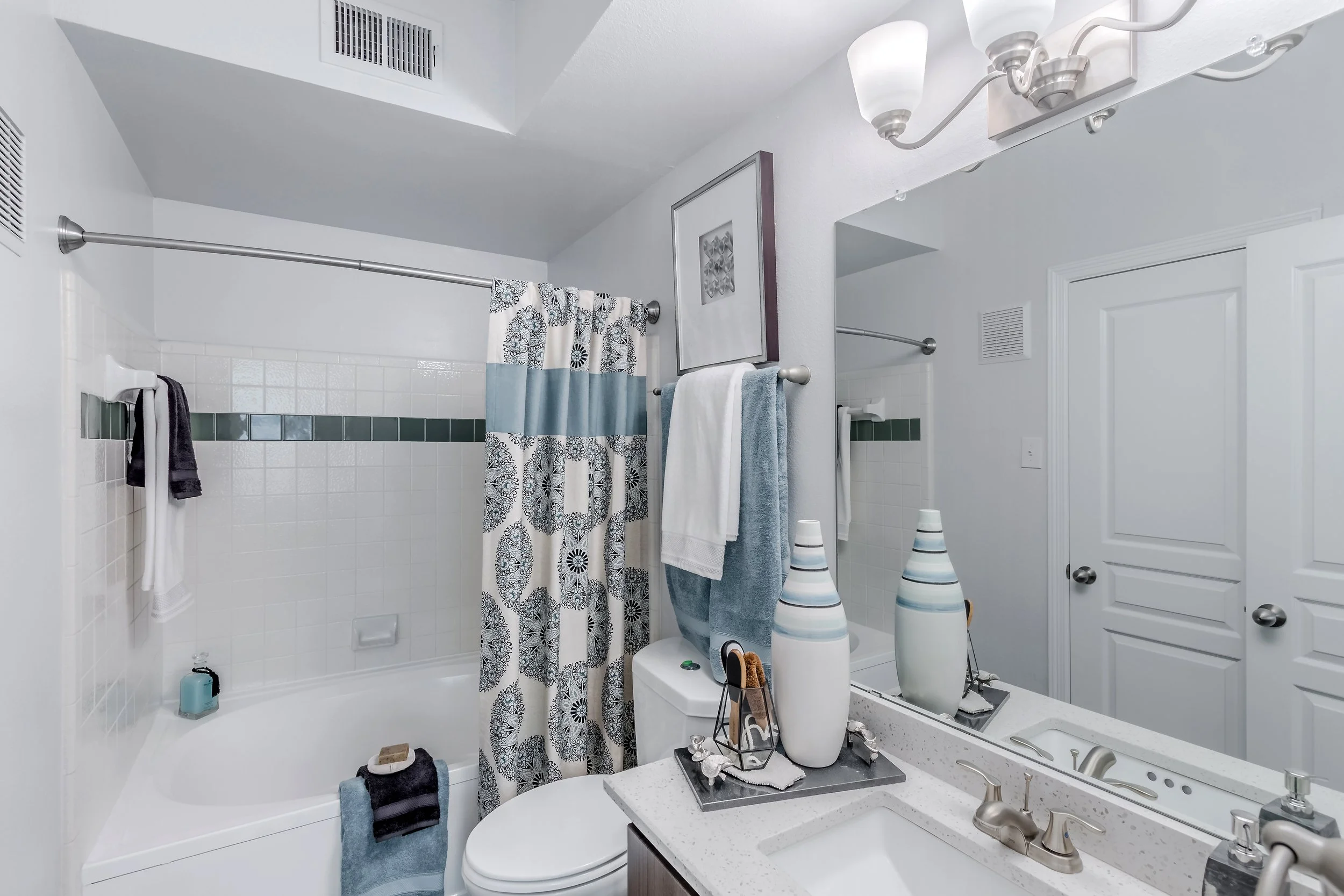 A bathroom with a bathtub and shower, a sink with a mirror, and various decorative items including vases and towels, with white walls and a ceiling vent.