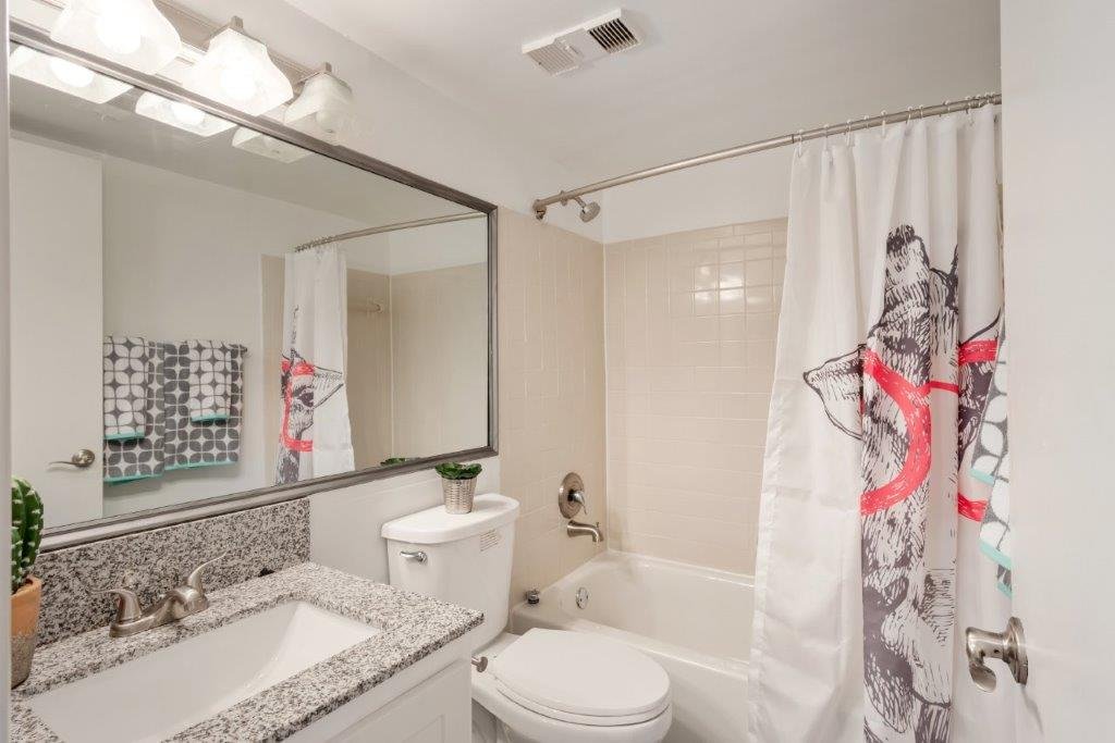 Bathroom with granite countertop sink, large mirror, small plant, toilet, bathtub with shower, decorative curtain, and towel rack with towels.