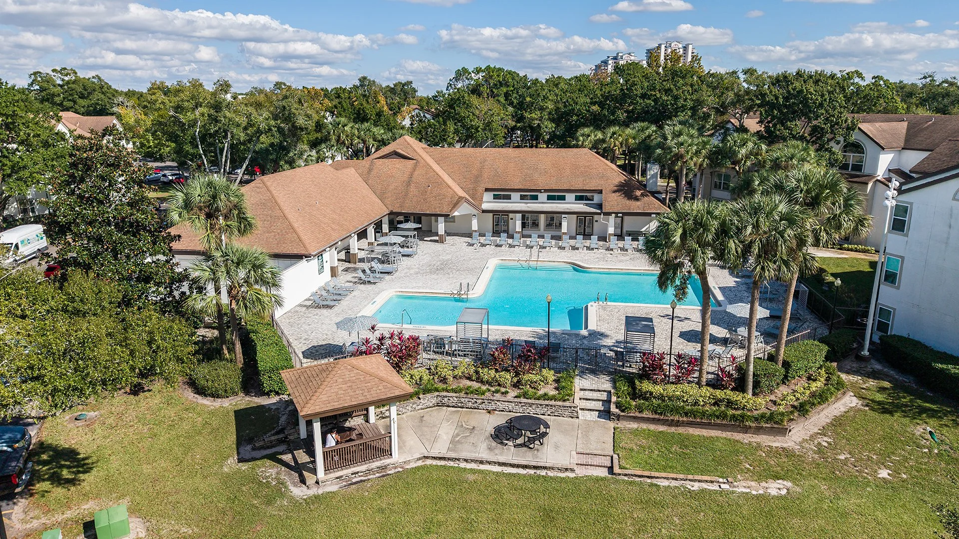 Aerial view of a community swimming pool area surrounded by trees, with a pavilion, lounge chairs, and a landscaped garden, in a residential complex during daytime.