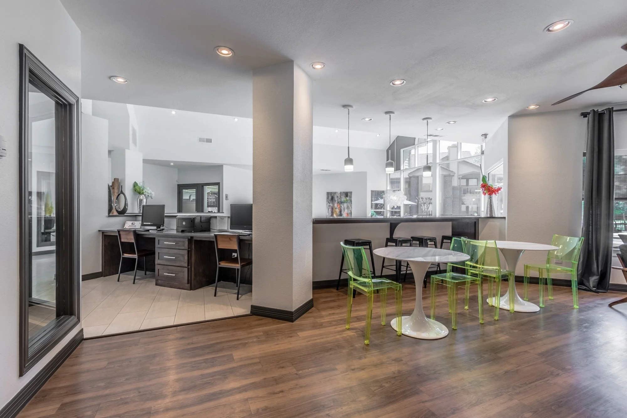 Interior of a modern lobby or café with wooden flooring, white walls, and large windows. There are green transparent chairs around white tables, a bar area with stools, and a reception desk with computer monitors.