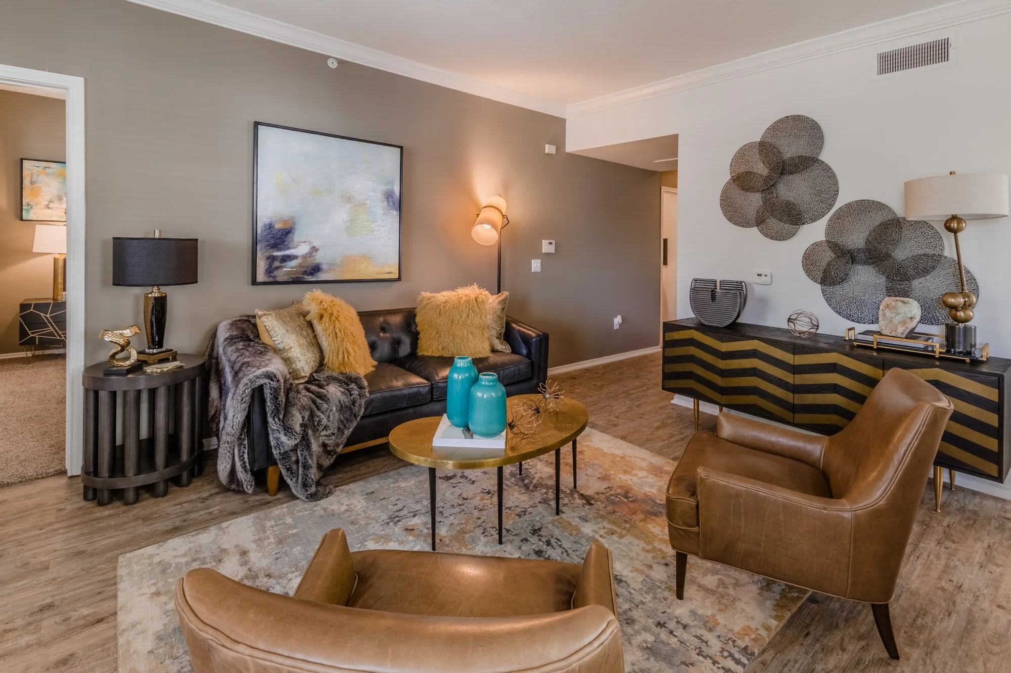 Living room with a gray wall, black leather sofa with yellow and gray pillows, a round golden coffee table with blue vases, two tan leather chairs, a black and gold sideboard, decorative wall art, table lamps, and a patterned rug.