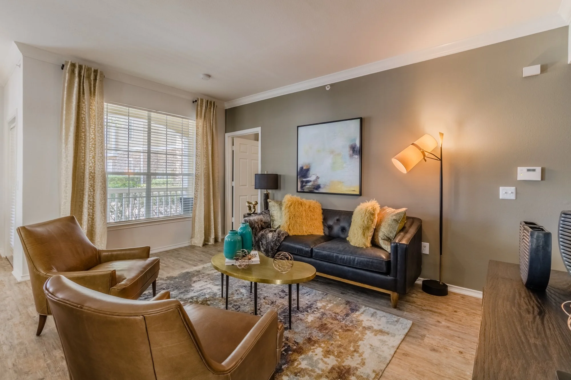 Living room with two brown leather armchairs, a black sofa with multiple fluffy pillows, a round coffee table with decorative vases, a rug, a large window with gold curtains, and wall art, lamp, and other furniture.