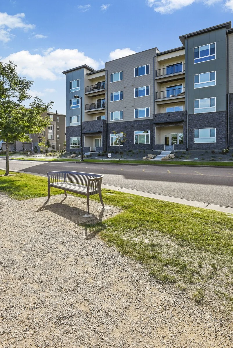 Modern apartment building with multiple balconies, a tree, a bench, and a clear blue sky.