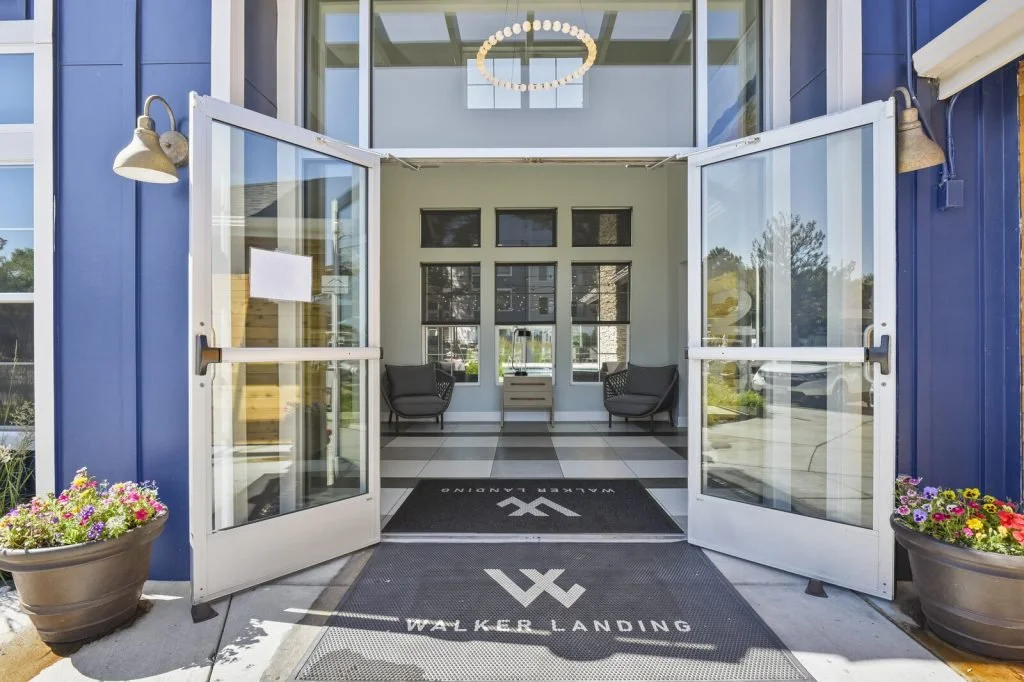 Open glass doors at the entrance of a building with a modern interior, two black chairs, potted flowers outside, and a black doormat with the logo and text "Walker Landing".