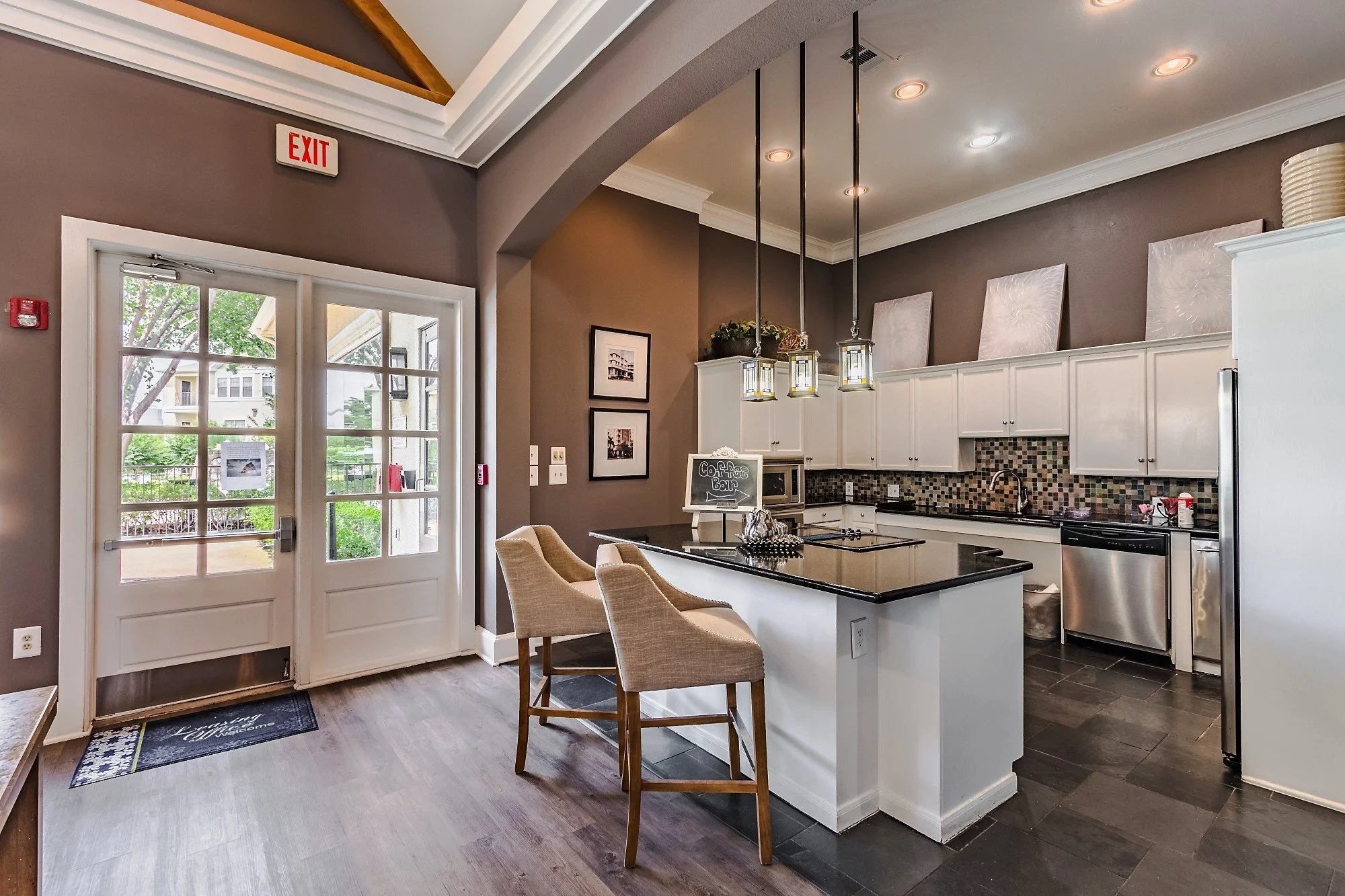 Kitchen with white cabinets, black countertops, and a mosaic tile backsplash. Two chairs are at a breakfast bar, and there is a dark tiled floor. Double glass doors lead outside, and framed pictures adorn the wall.