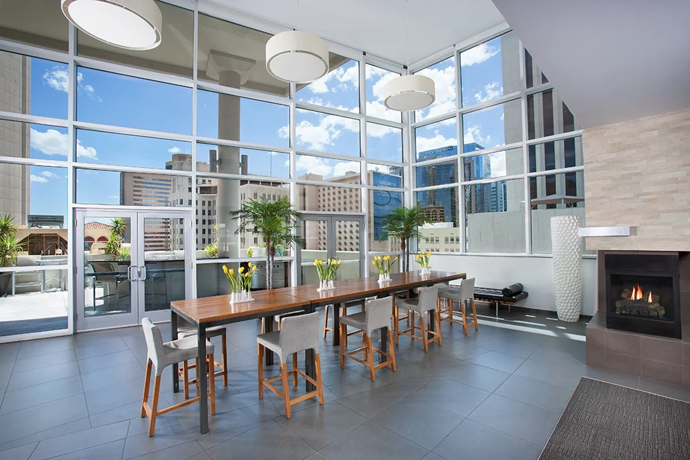 Bright indoor dining area with large floor-to-ceiling windows showing city buildings, a long wooden table with nine chairs, vases with yellow flowers, two potted green plants, a fireplace, and modern hanging lights.