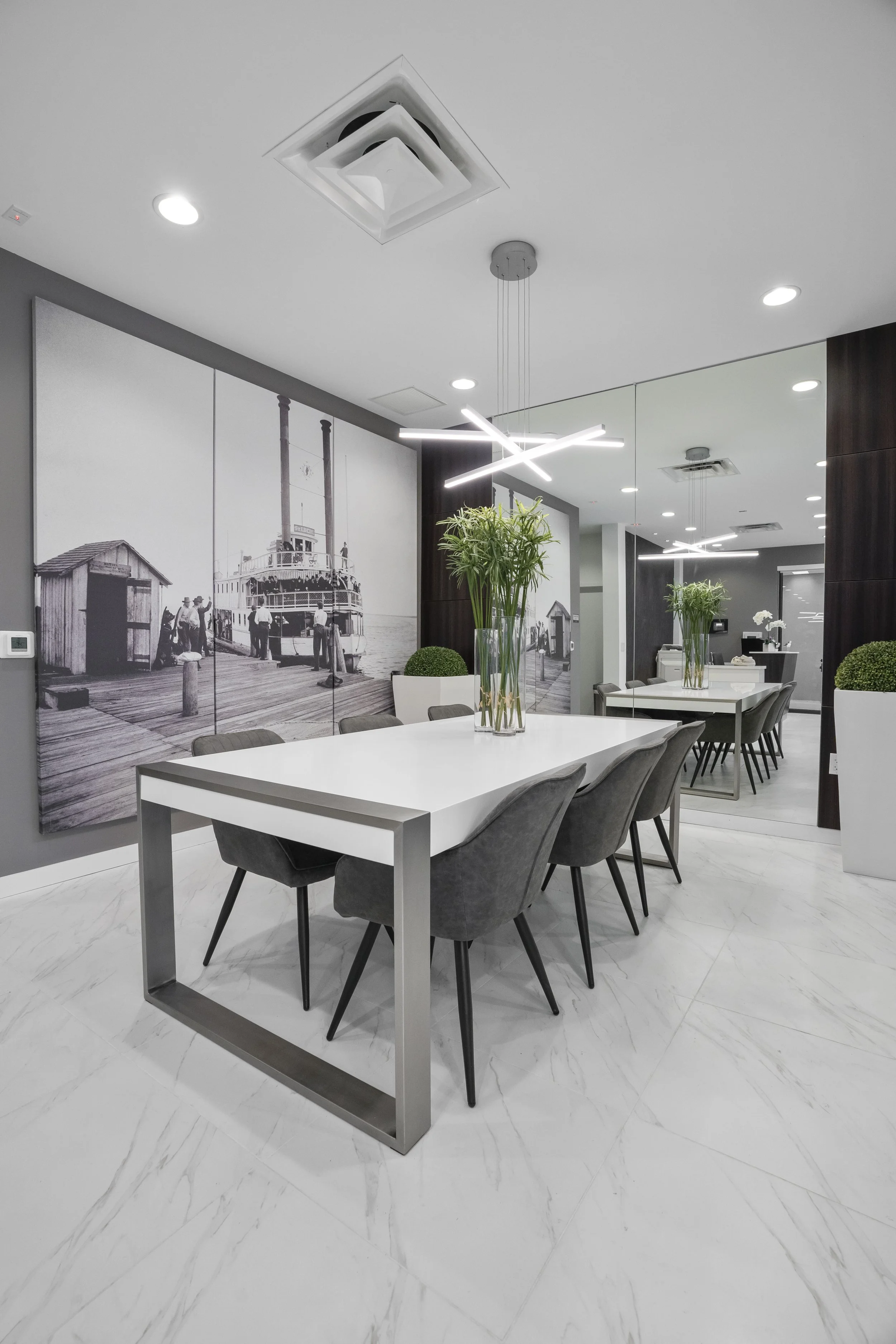 Modern dining room with white table, gray chairs, large black-and-white photograph of a seaside dock scene, and white planters with green plants.