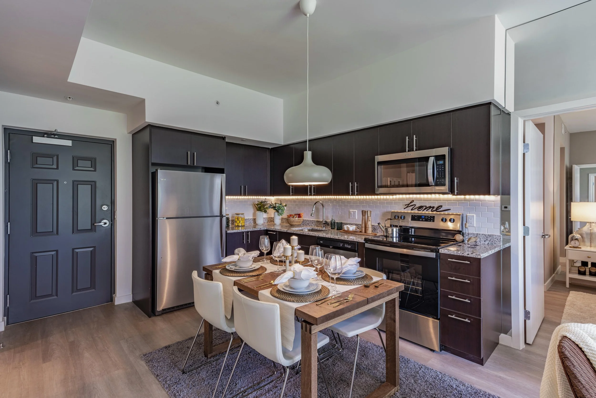 Modern kitchen with dark cabinets, stainless steel refrigerator, microwave, and oven, white subway tile backsplash, granite countertop, dining table set for four with white chairs, candles, and tableware, pendant light, and minimalist decor.