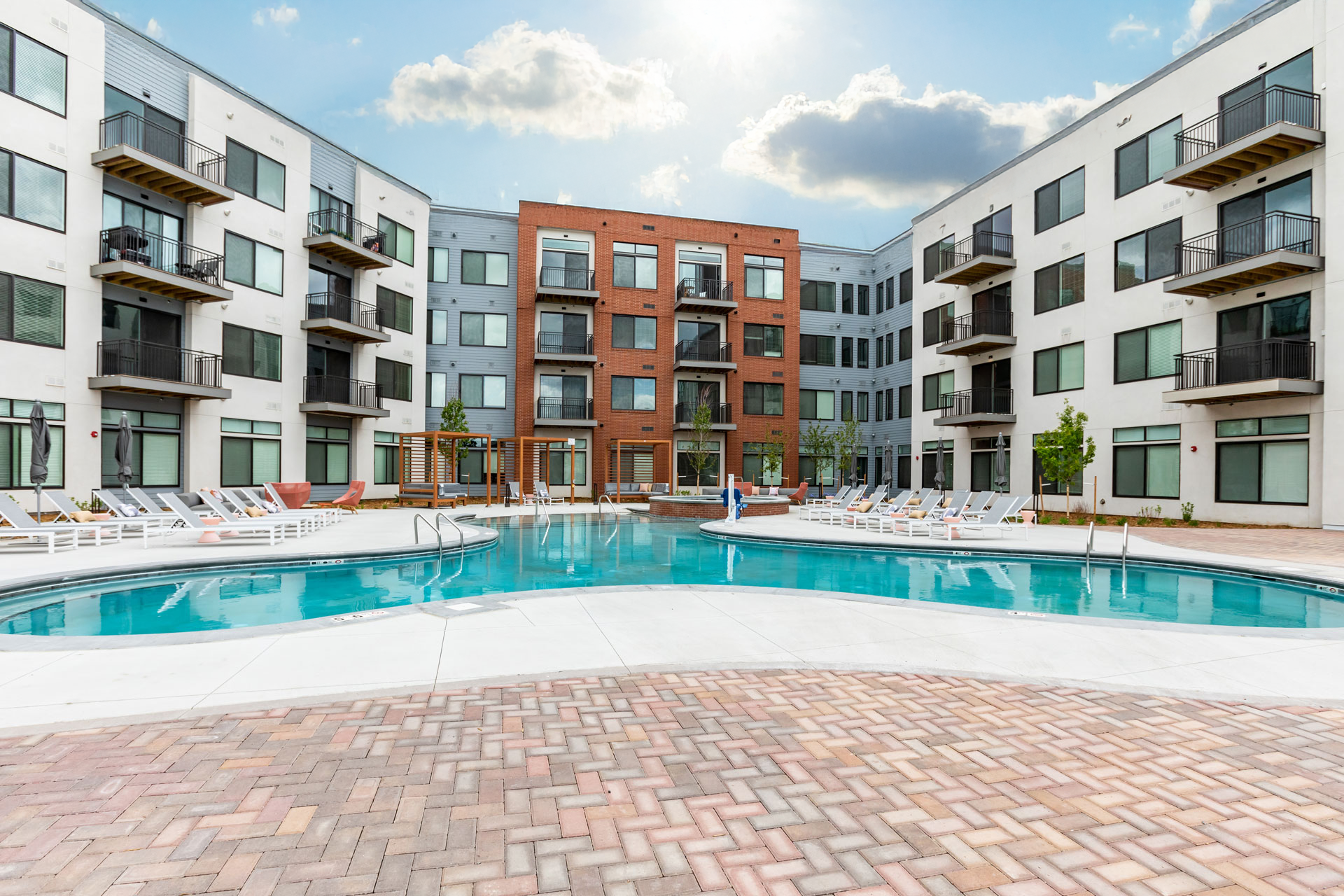 Image of a modern apartment complex with a swimming pool in the courtyard, surrounded by lounge chairs, small trees, and shaded seating areas.