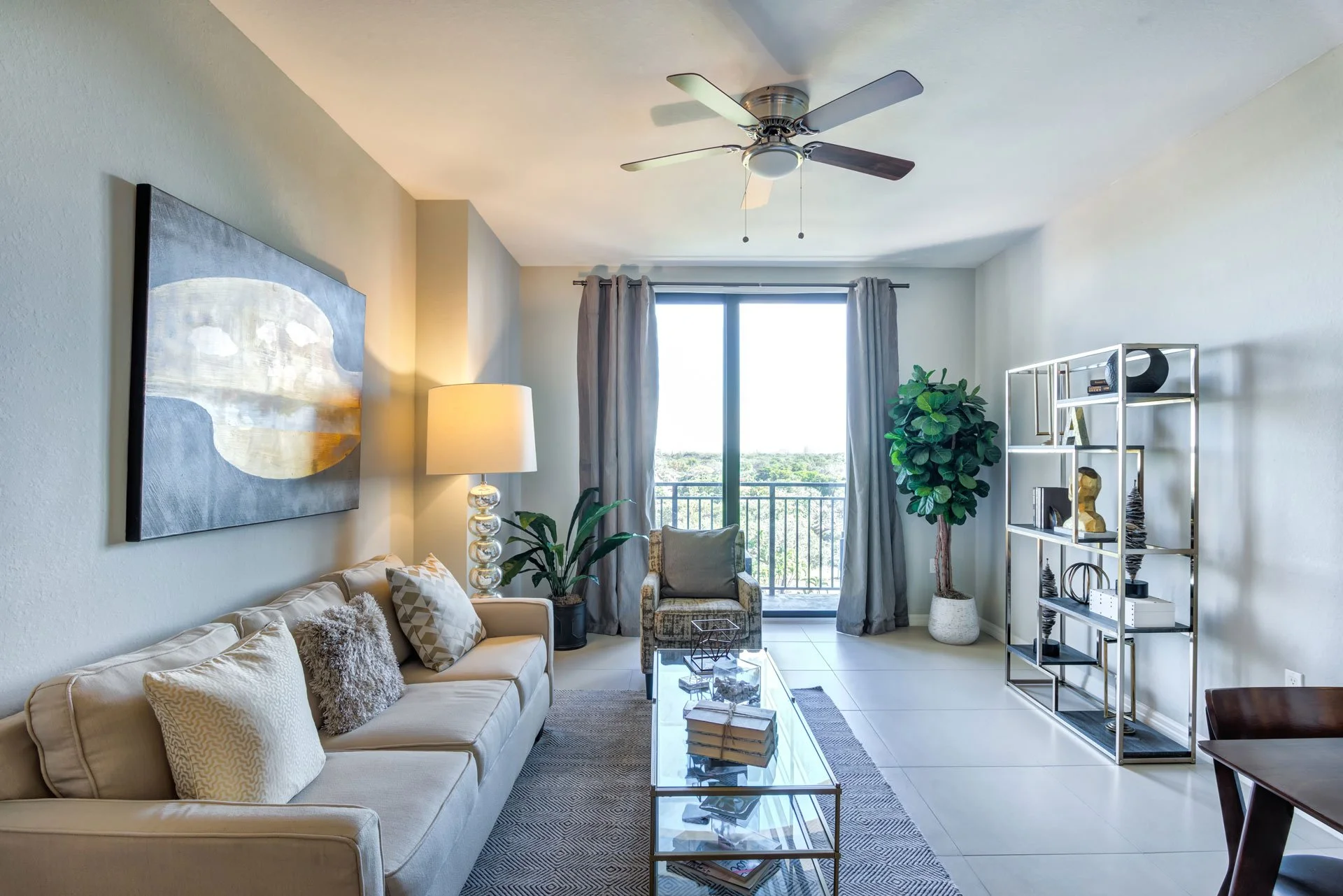 Living room with a cream sofa, a glass coffee table, a patterned armchair, and a metal bookshelf. Large window with gray curtains, potted plant, ceiling fan, and artwork on the wall.