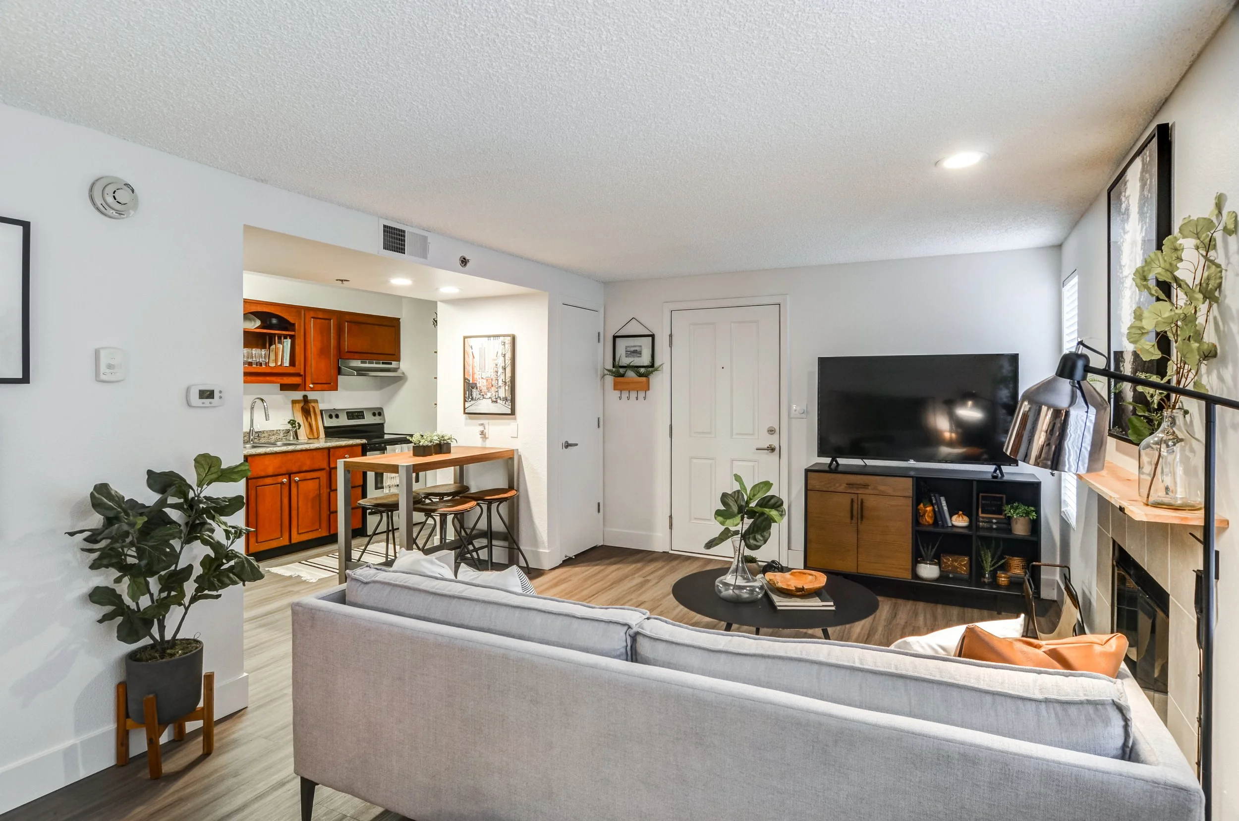 Open-concept living room and kitchen with white walls, dark wood floors, and contemporary furniture. The living room features a gray sofa, a black coffee table, and a flat-screen TV on a wooden stand. The kitchen has wooden cabinets, a small counter 
