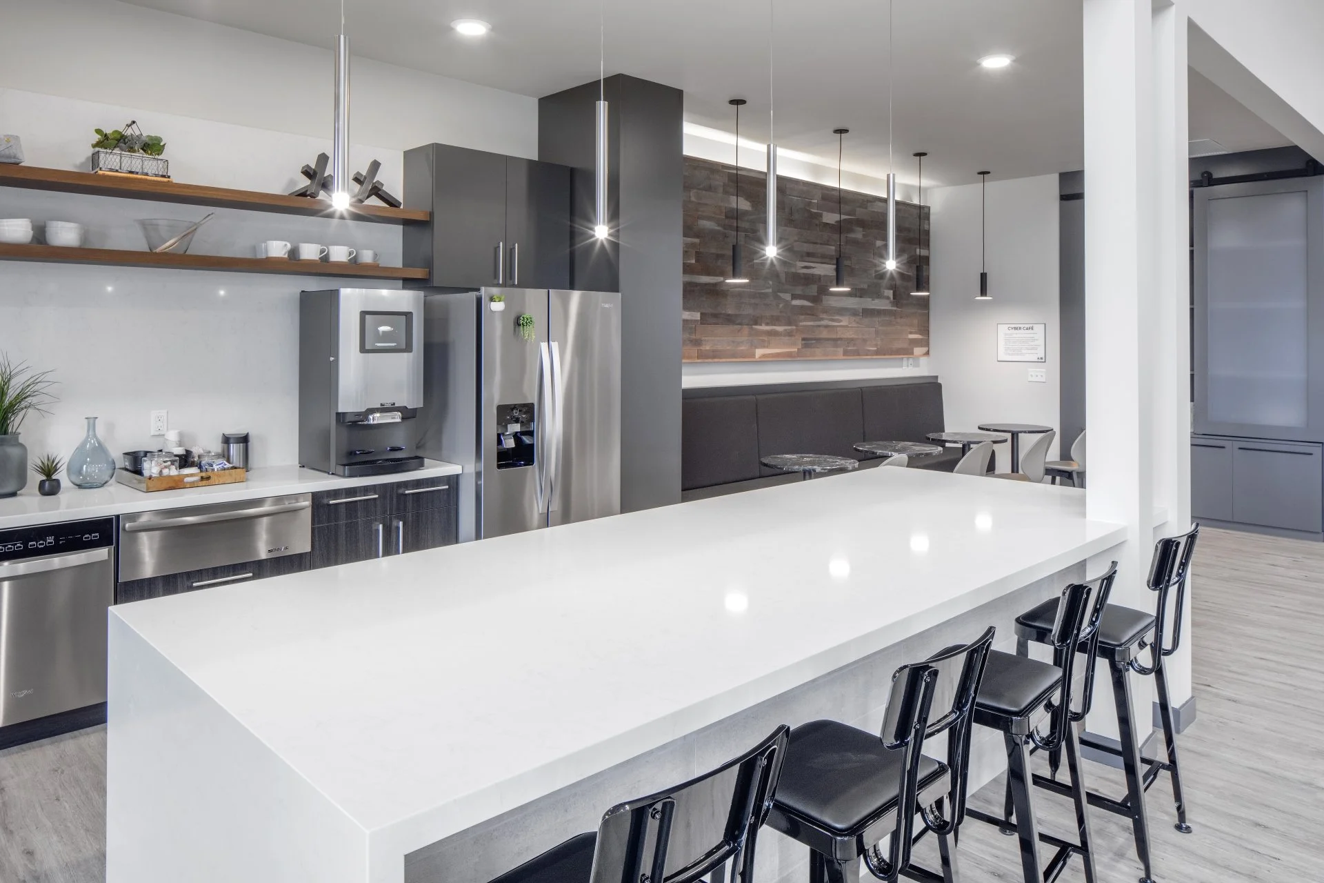 Modern kitchen with white island counter, black chairs, stainless steel refrigerator, coffee machine on countertop, open shelving, wood accent wall, and small dining area with tables and chairs.