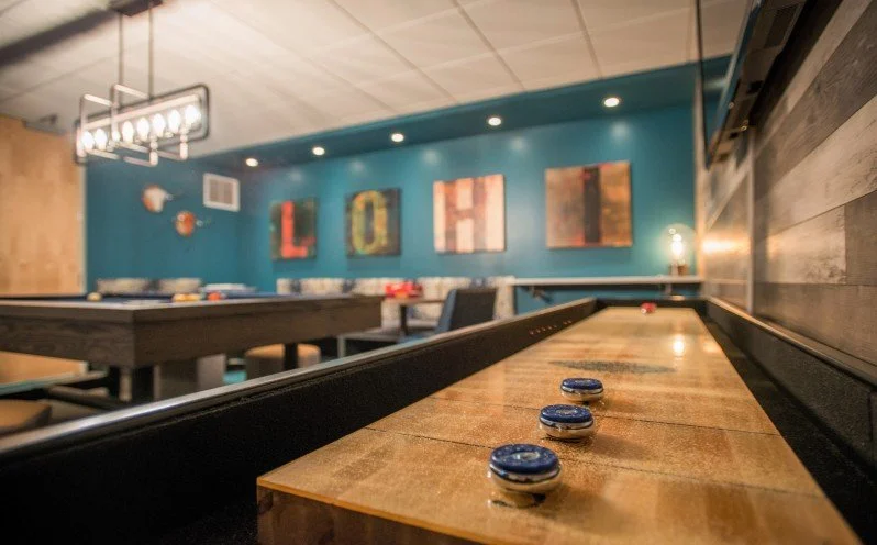Close-up of a shuffleboard table with pucks, inside a decorated game room with a blue wall, colorful artwork, and set up for entertainment.