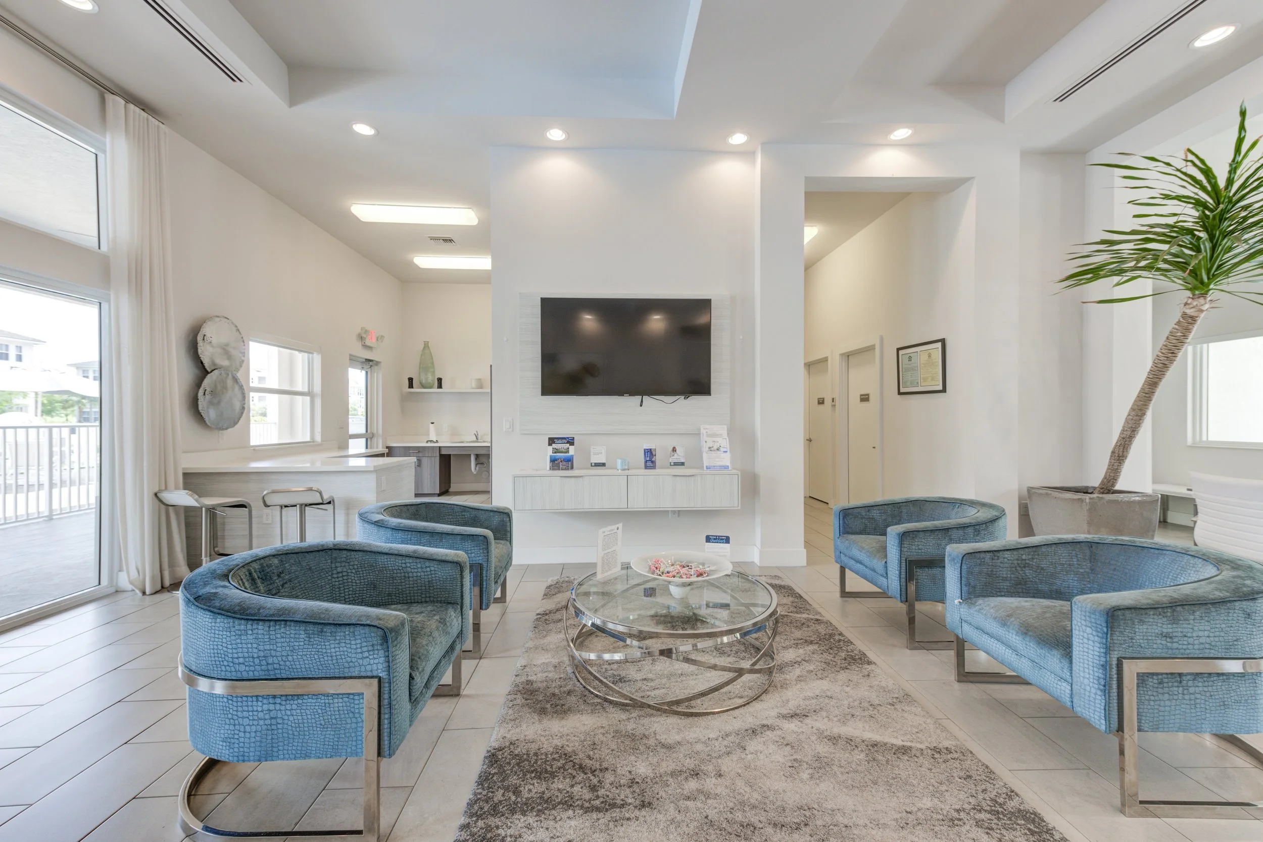 Modern hotel lobby with blue velvet armchairs, a glass coffee table, large window, TV on the wall, and decorative plants.