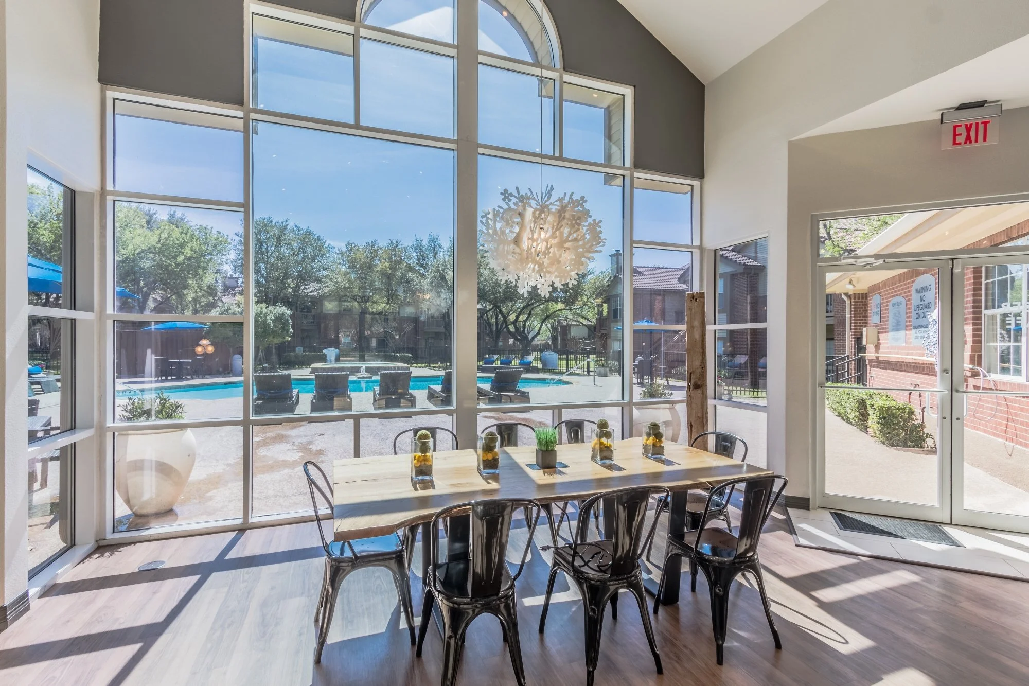 Indoor dining area with a wooden table and black chairs, decorated with small vases containing yellow flowers and green plants, overlooking a pool outside through large windows, with a modern chandelier hanging from the ceiling and an exit door on th