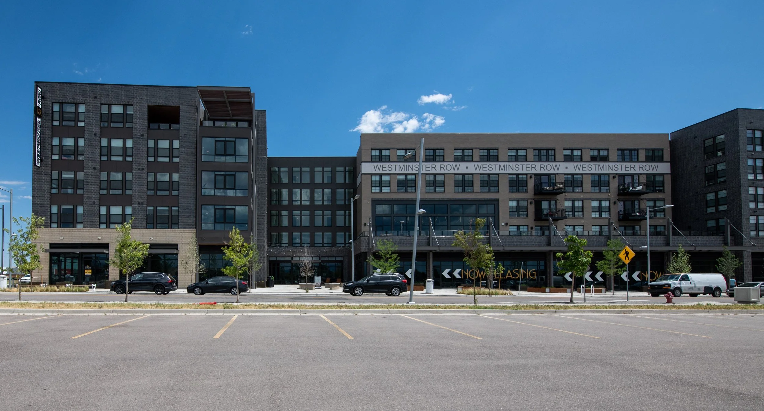 Modern multi-story residential or commercial building with glass windows, trees lining the sidewalk, and a parking lot in front on a clear, sunny day.