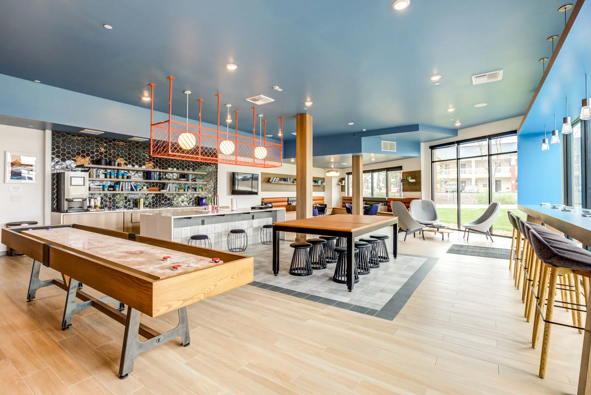 Modern hotel lounge with a ping pong table, bar area, and seating with chairs near large windows showing an outdoor grassy area.