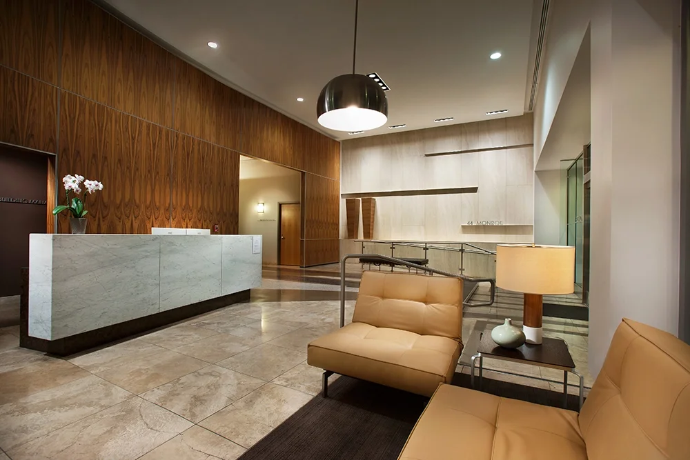 Modern hotel lobby with wooden wall paneling, marble reception desk, beige seating, a floor lamp, and orchid plant on the desk.