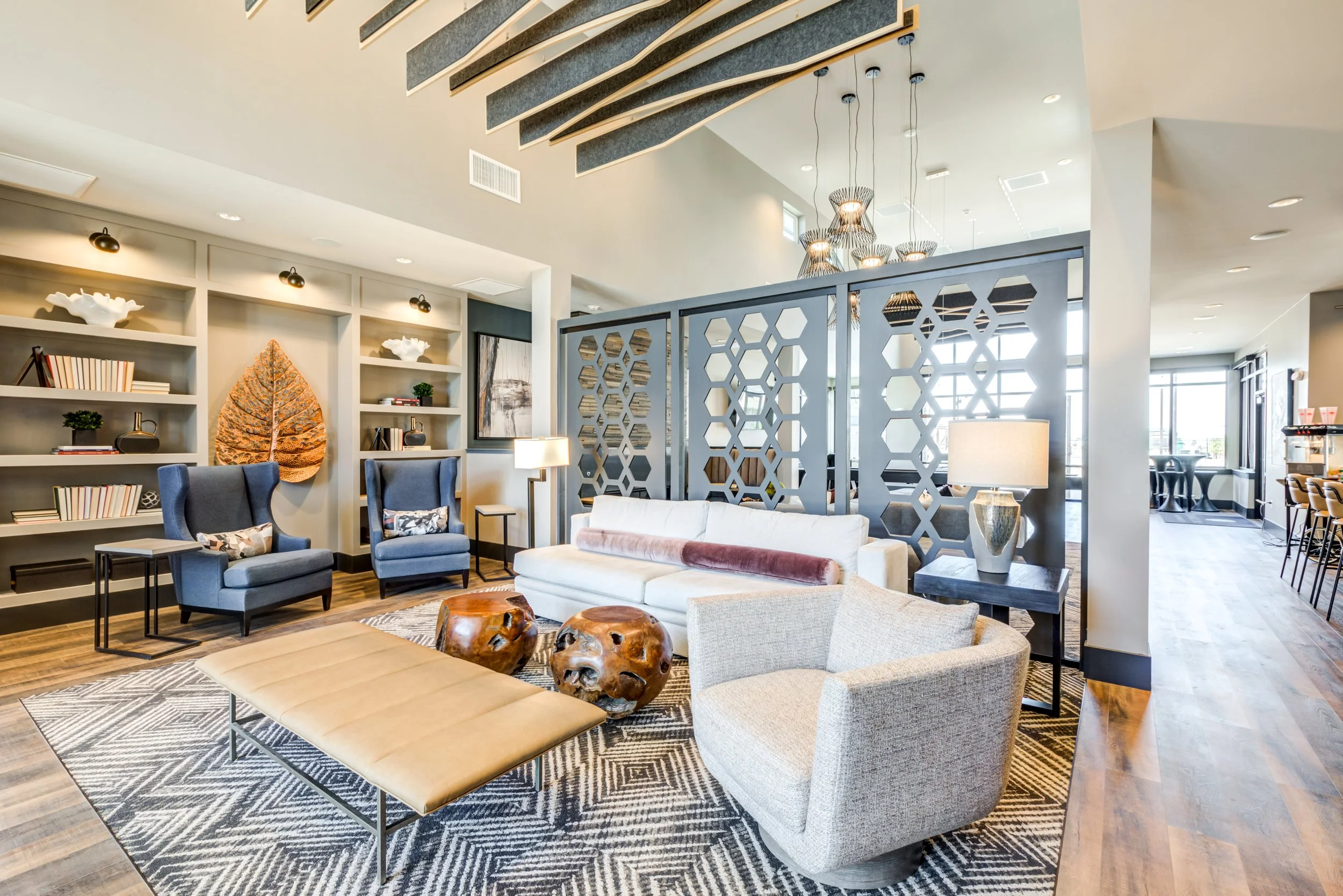 Modern hotel lobby with seating area, including white sofa, beige armchair, and blue armchairs, with decorative wooden stools, bookshelves, and a partition with hexagonal cutouts, all illuminated by natural light and overhead lighting.