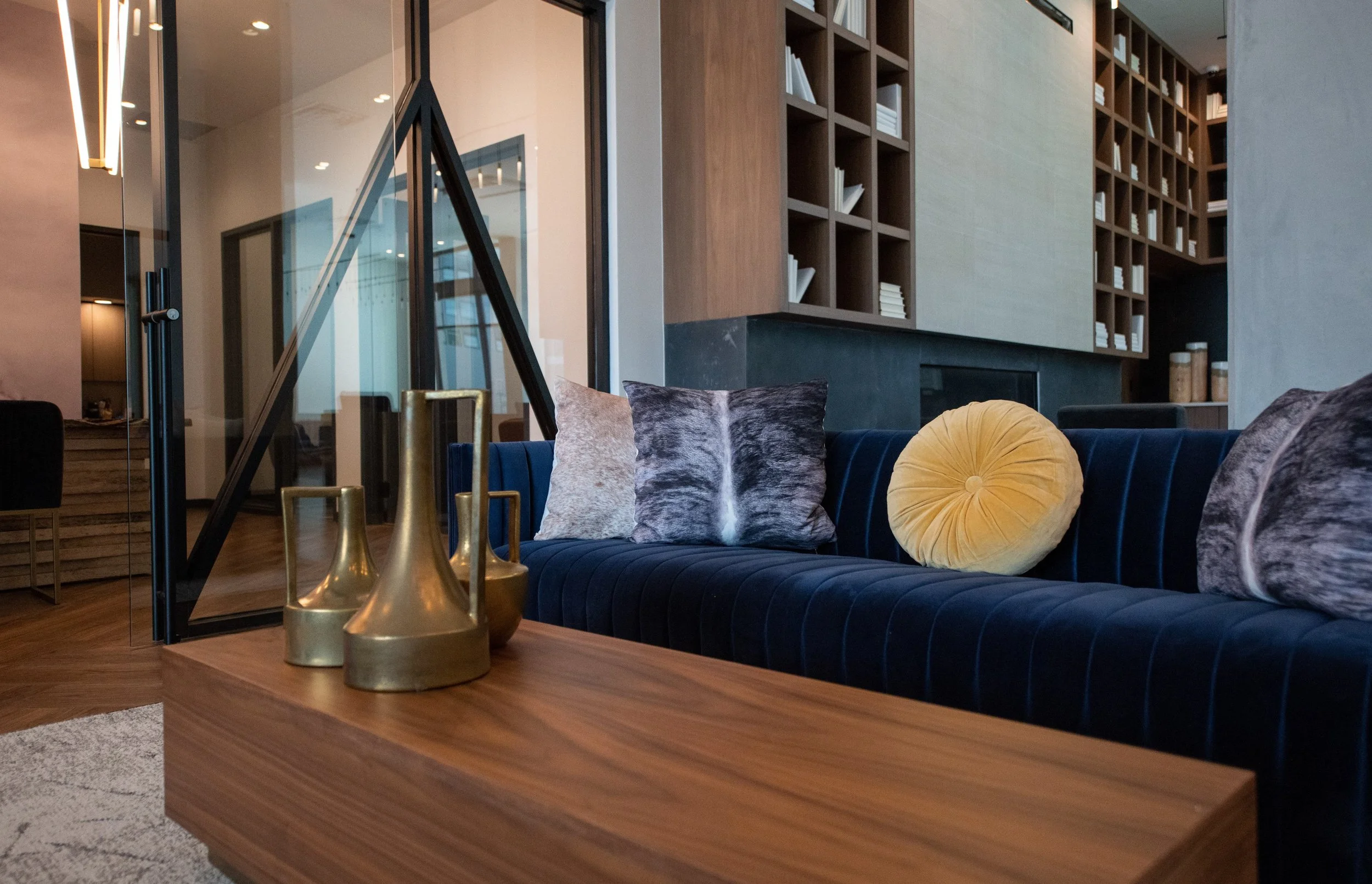 Modern living room with a navy velvet sofa, decorative pillows, a wooden coffee table with brass vases, and a background of wooden bookshelves and glass doors.