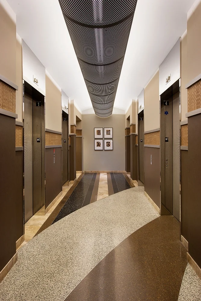 Hotel elevator corridor with six elevators, artwork on back wall, patterned ceiling, and tiled floor.