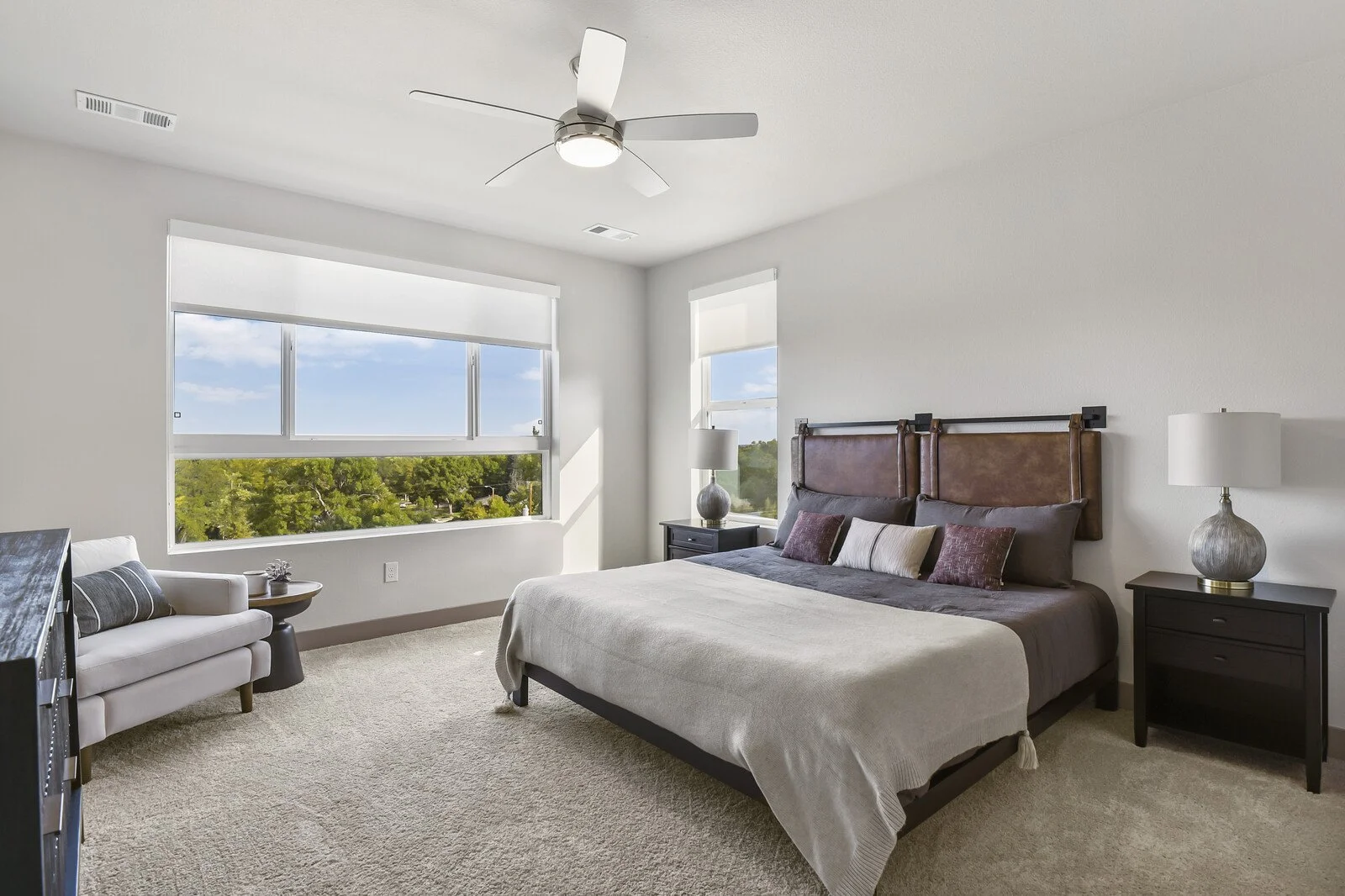 A bedroom with a large bed, nightstands with lamps, a white armchair, a ceiling fan, and large windows showing trees and a blue sky.