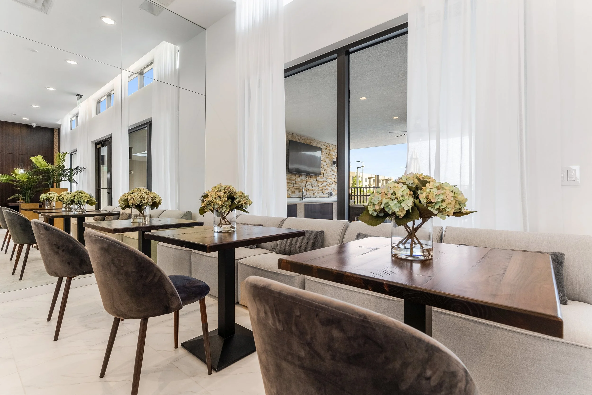 Interior of a modern dining area with wooden tables, velvet chairs, and vases of white flowers, alongside a large window with sheer white curtains.