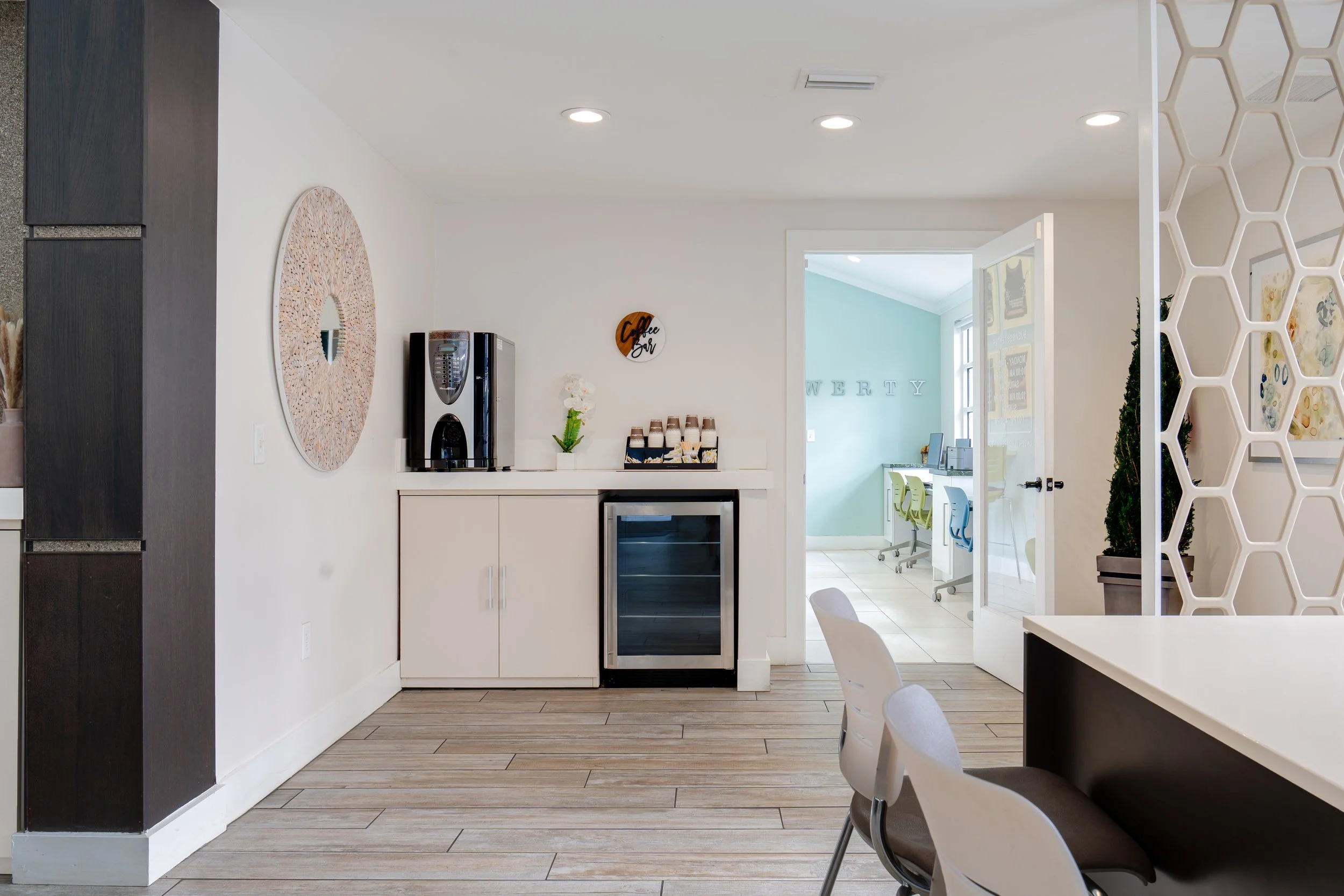 Interior view of a modern coffee service area with a coffee machine, small refrigerator, and decorative wall art, adjacent to an office area with desks and chairs.