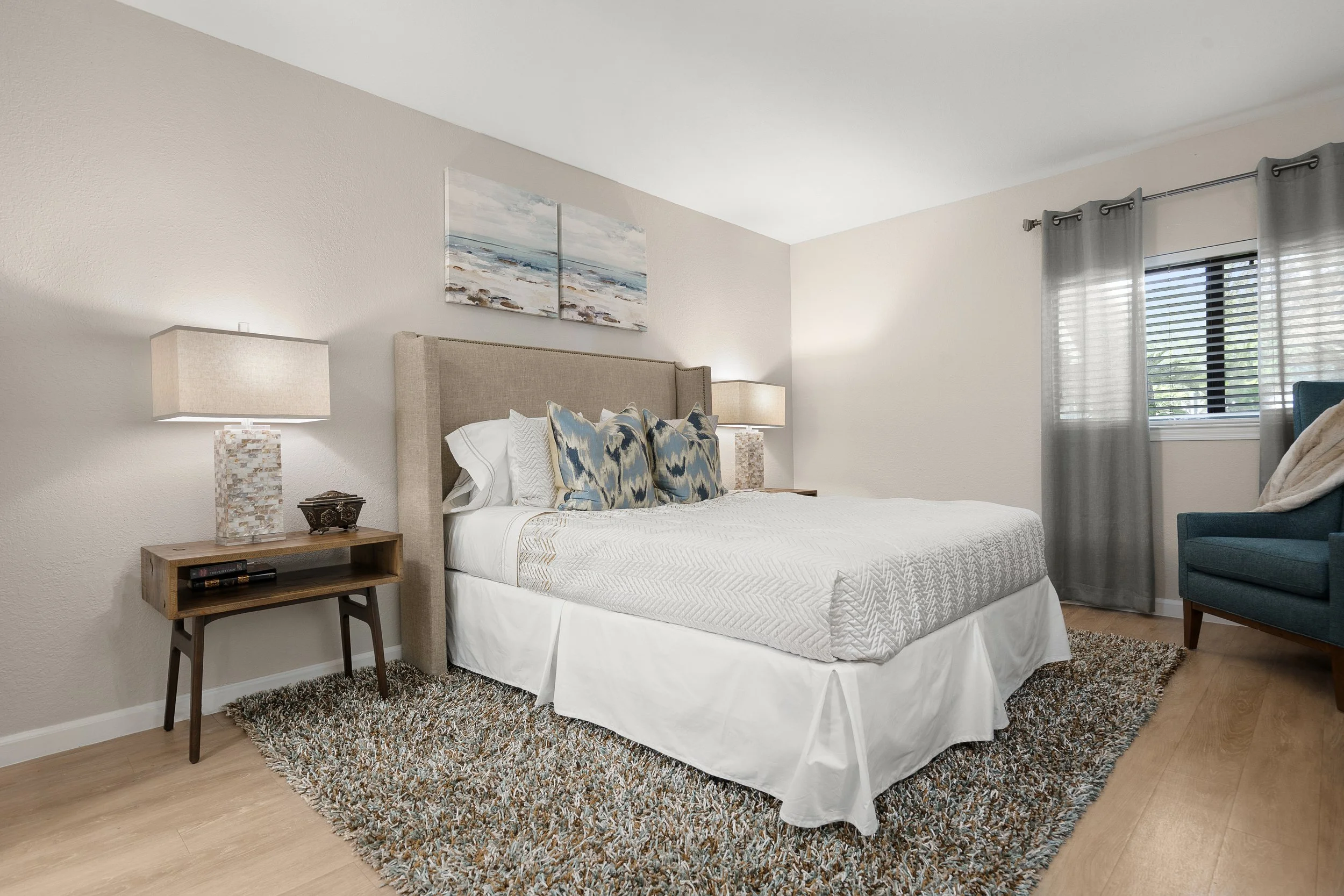 A bedroom with a large bed, beige headboard, white bedding, and several blue patterned pillows. There are two bedside tables with lamps, a rug on the wooden floor, a window with gray curtains, and a teal armchair with a blanket.