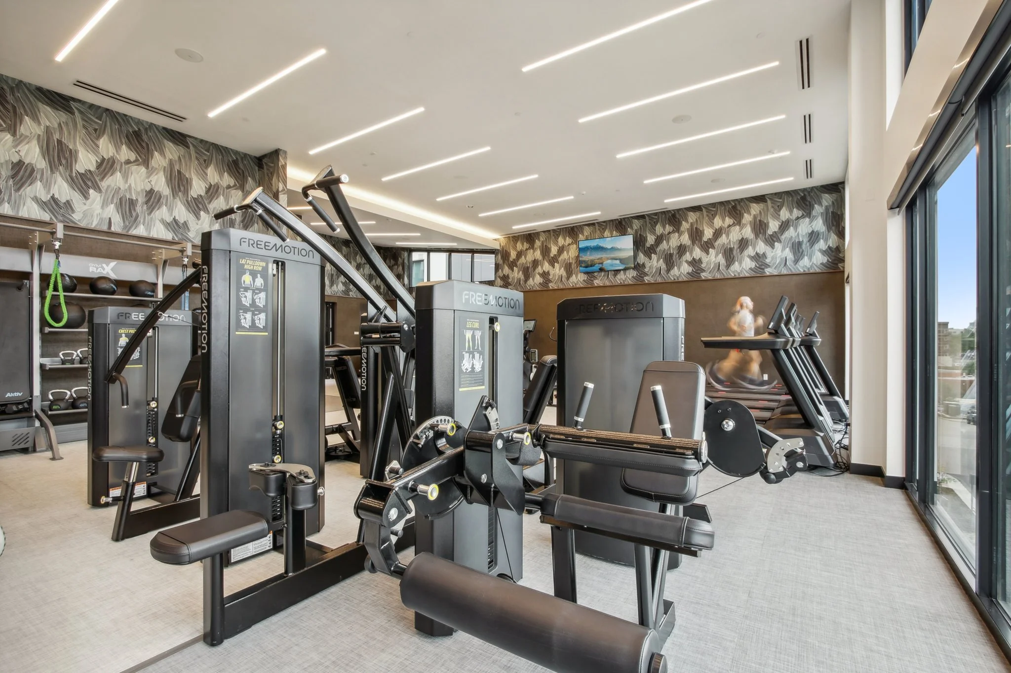 Modern gym with various black workout equipment, large windows, and a TV mounted on the wall.