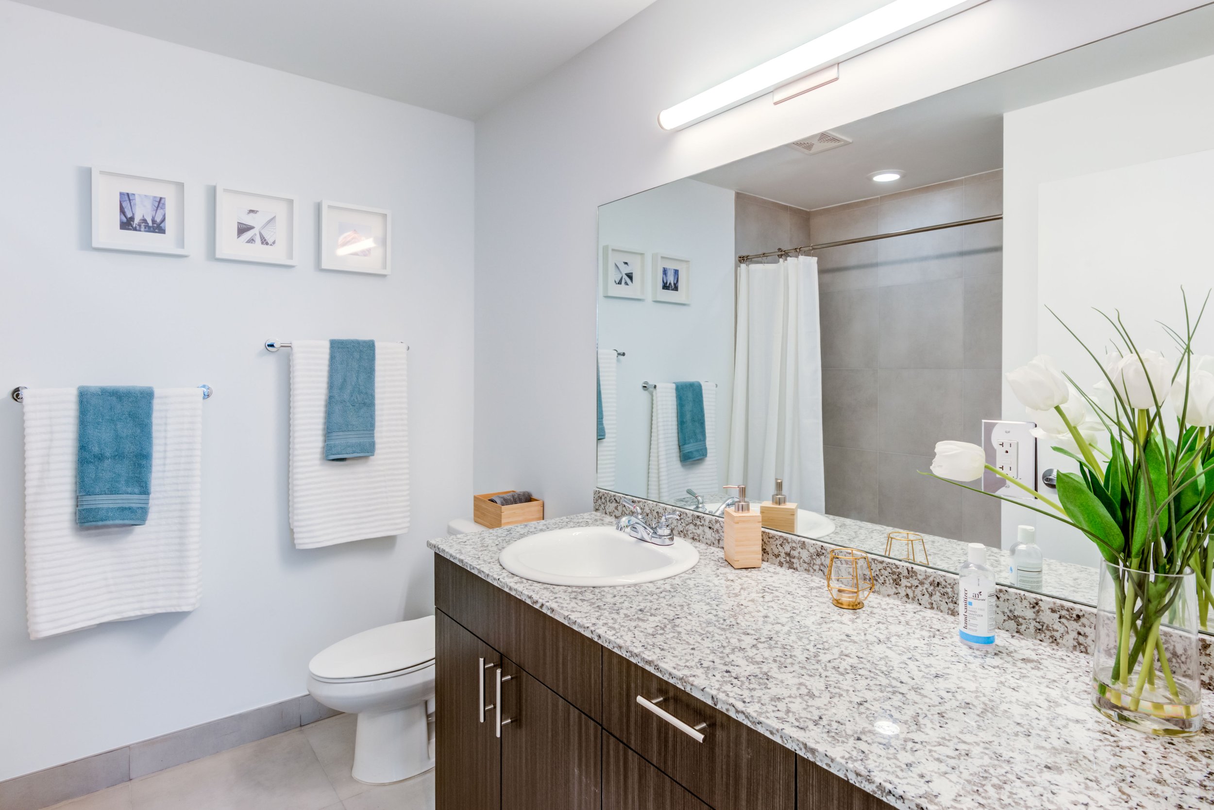 Modern bathroom with granite countertop, flowers in a glass vase, towels, framed pictures, beard care products, and a mirror reflecting a shower area with a white curtain.