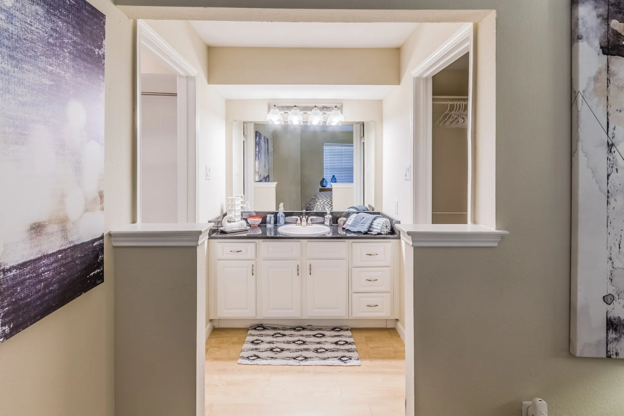 A bathroom with a single sink vanity with white cabinets, a dark granite countertop, a mirror, and four light bulbs above. The vanity is decorated with blue towels, a soap dispenser, and a small bowl, with a patterned rug on the wooden floor. The mir