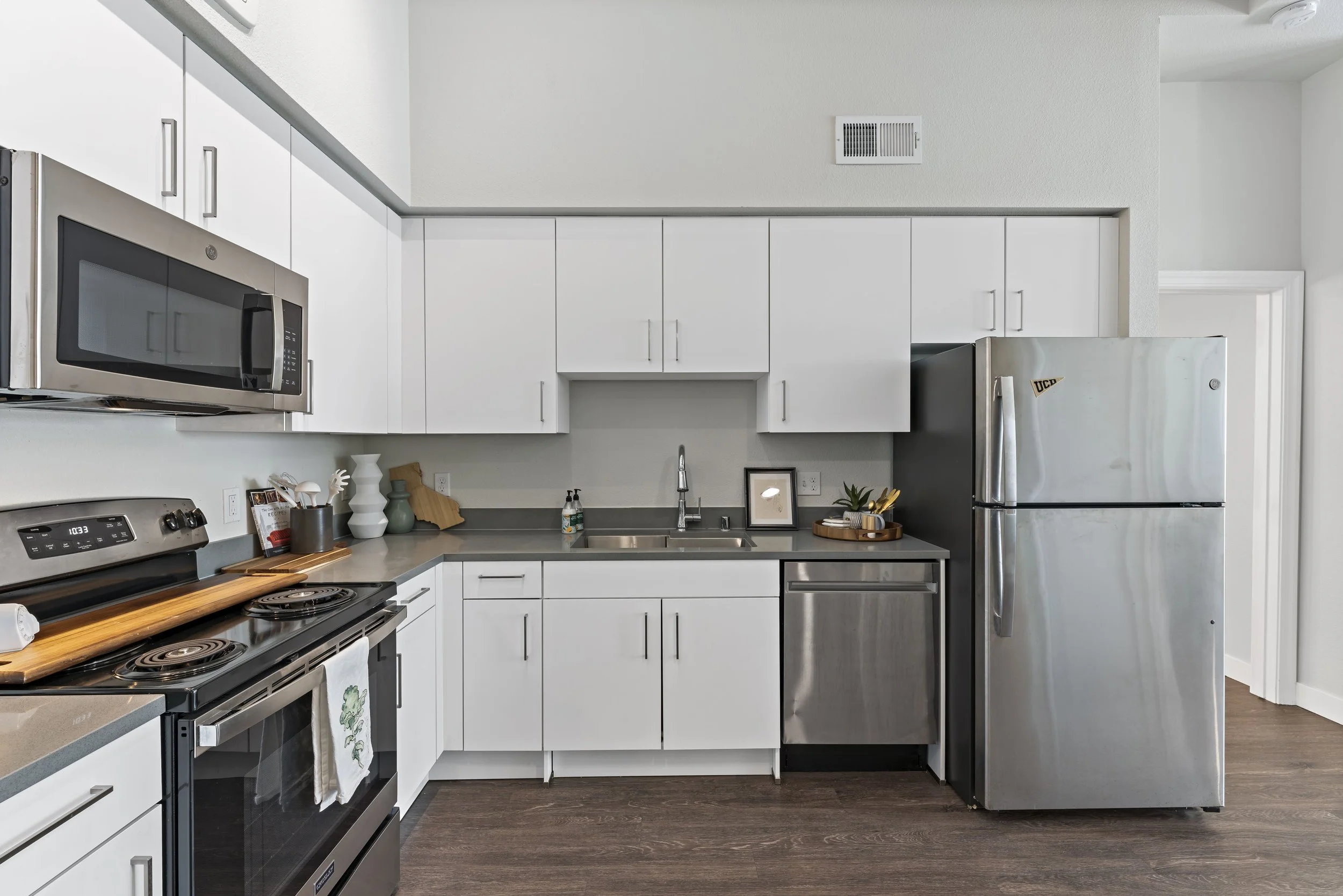Modern kitchen with white cabinets, stainless steel appliances including a microwave, oven, and refrigerator, gray countertops, and dark wood flooring.