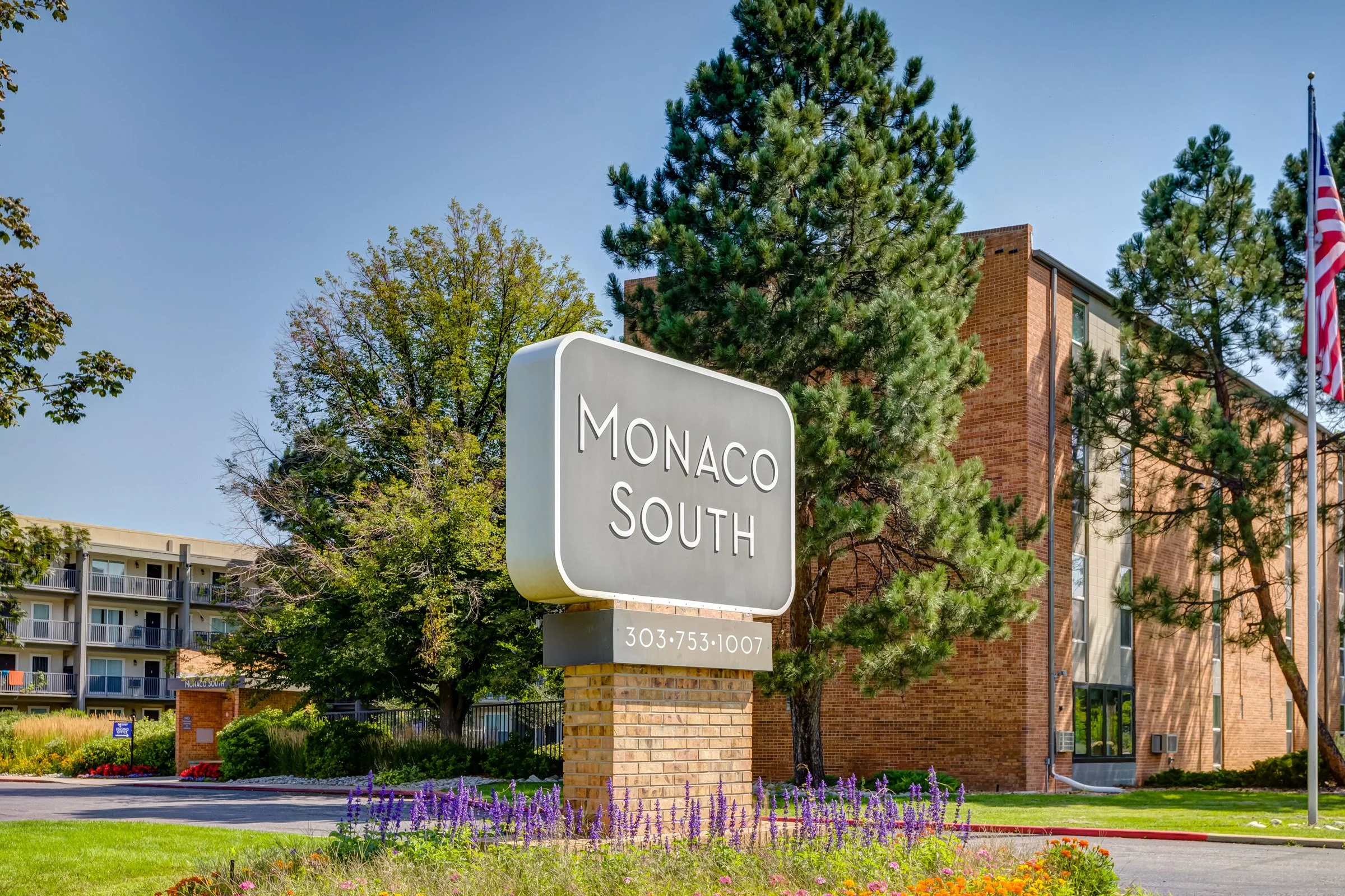 Sign for Monaco South apartment complex or neighborhood with a phone number, in front of landscaped area with flowers, trees, and a brick building, with added greenery and American flags.