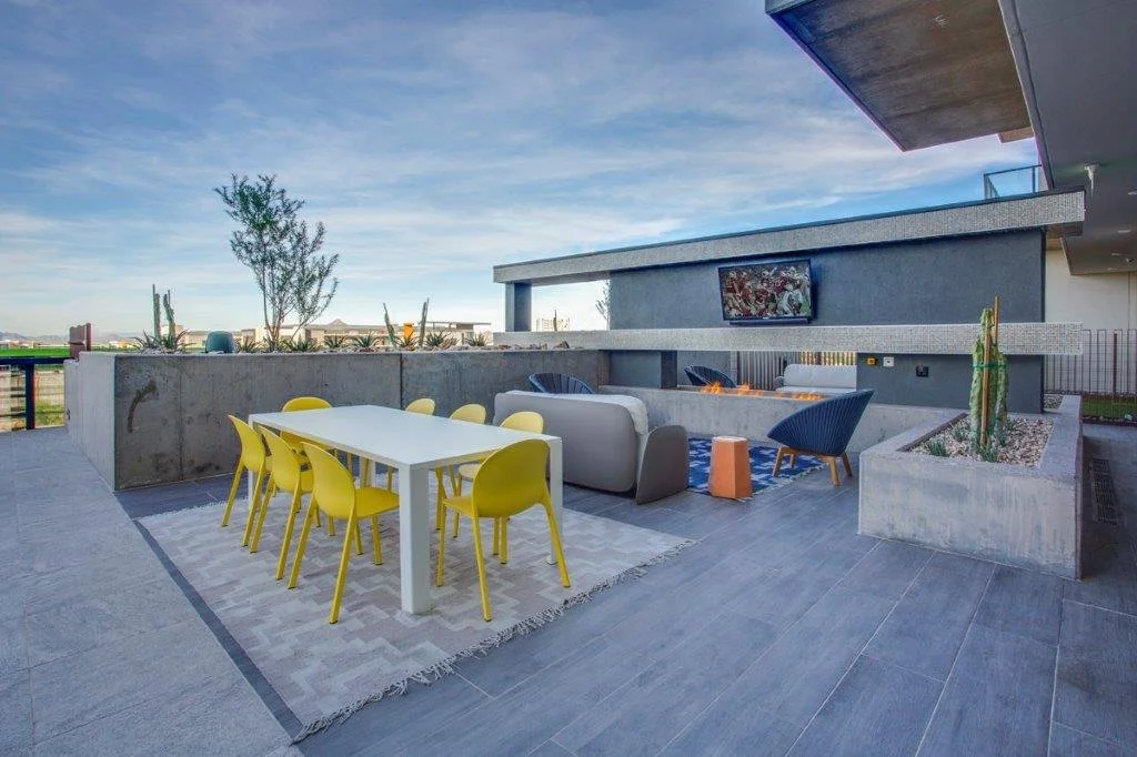 Outdoor rooftop terrace with a modern gray concrete wall, yellow dining chairs around a white table, a fireplace with lounge chairs, a wall-mounted TV, and potted desert plants, under a partly cloudy sky.